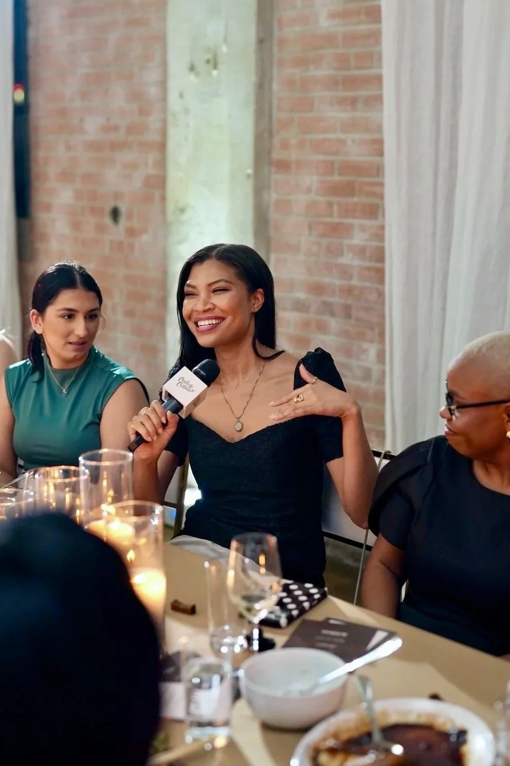 Woman in black dress speaking into a microphone at a gathering, seated at a table with other women and food and drinks, in a room with brick and white walls.