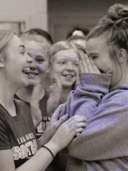 A group of girls at school, with one girl covering her face and others smiling and laughing.