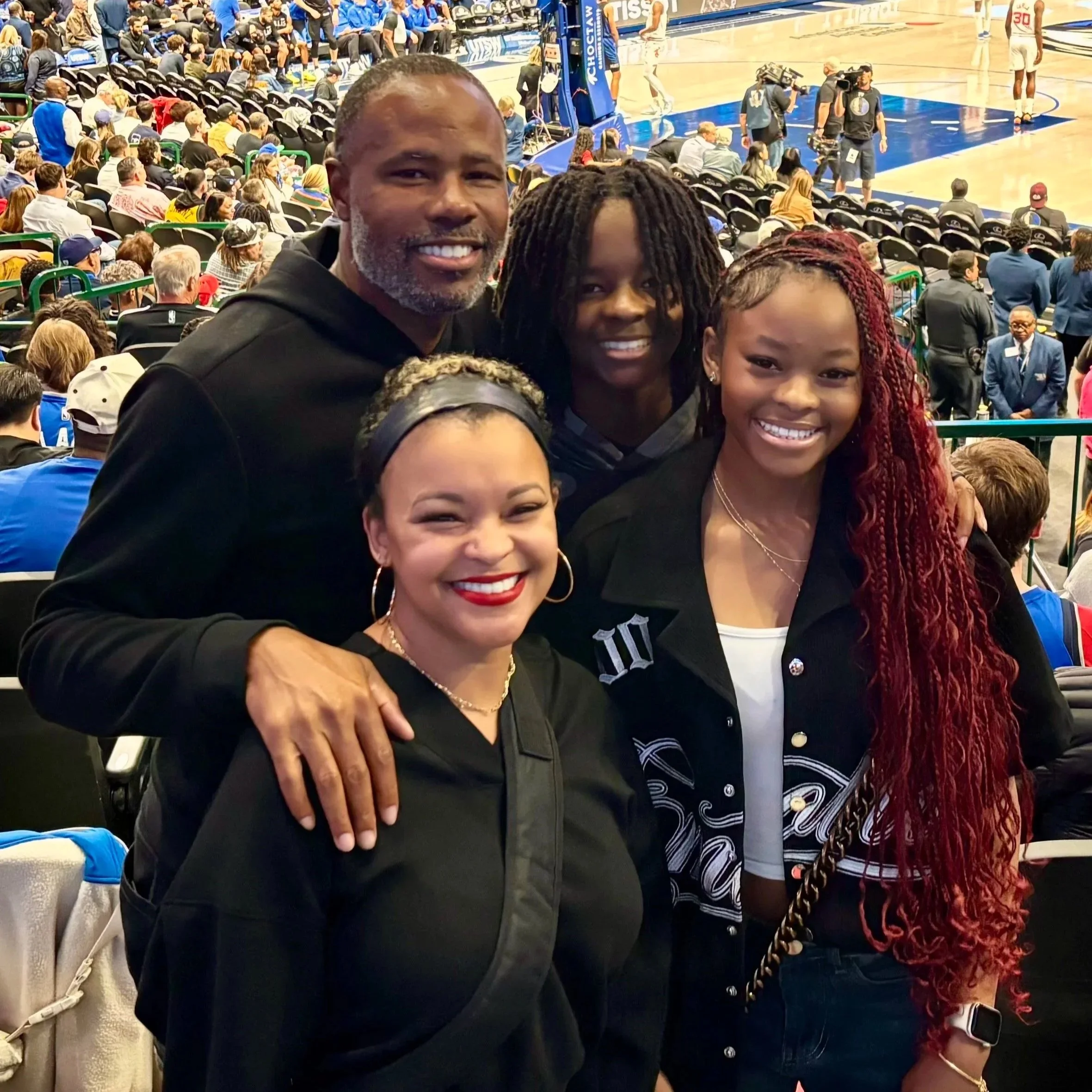 A group of people sitting courtside during a basketball game with some flashing cameras. The focus is on three individuals in the foreground, smiling, with one man and two women. The background shows a crowded arena with spectators and staff.