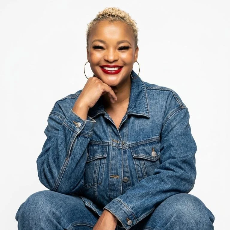 Smiling woman with blonde curly hair, red lipstick, hoop earrings, wearing a denim jacket and jeans, sitting with one hand resting on her chin against a plain white background.
