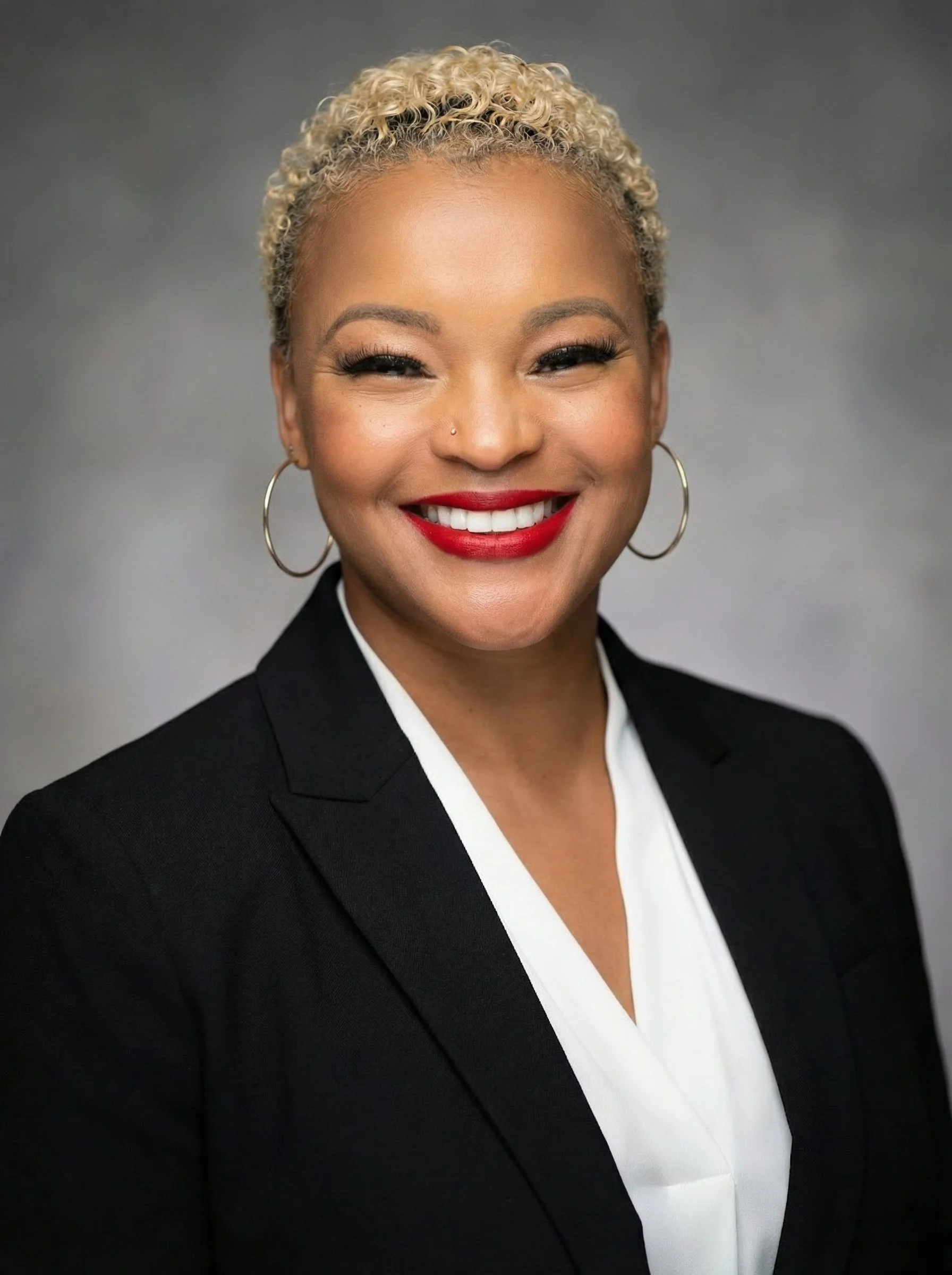 A photo of Crystal Patton smiling with blonde curly hair, wearing hoop earrings and a black blazer with a white shirt, against a gray background.