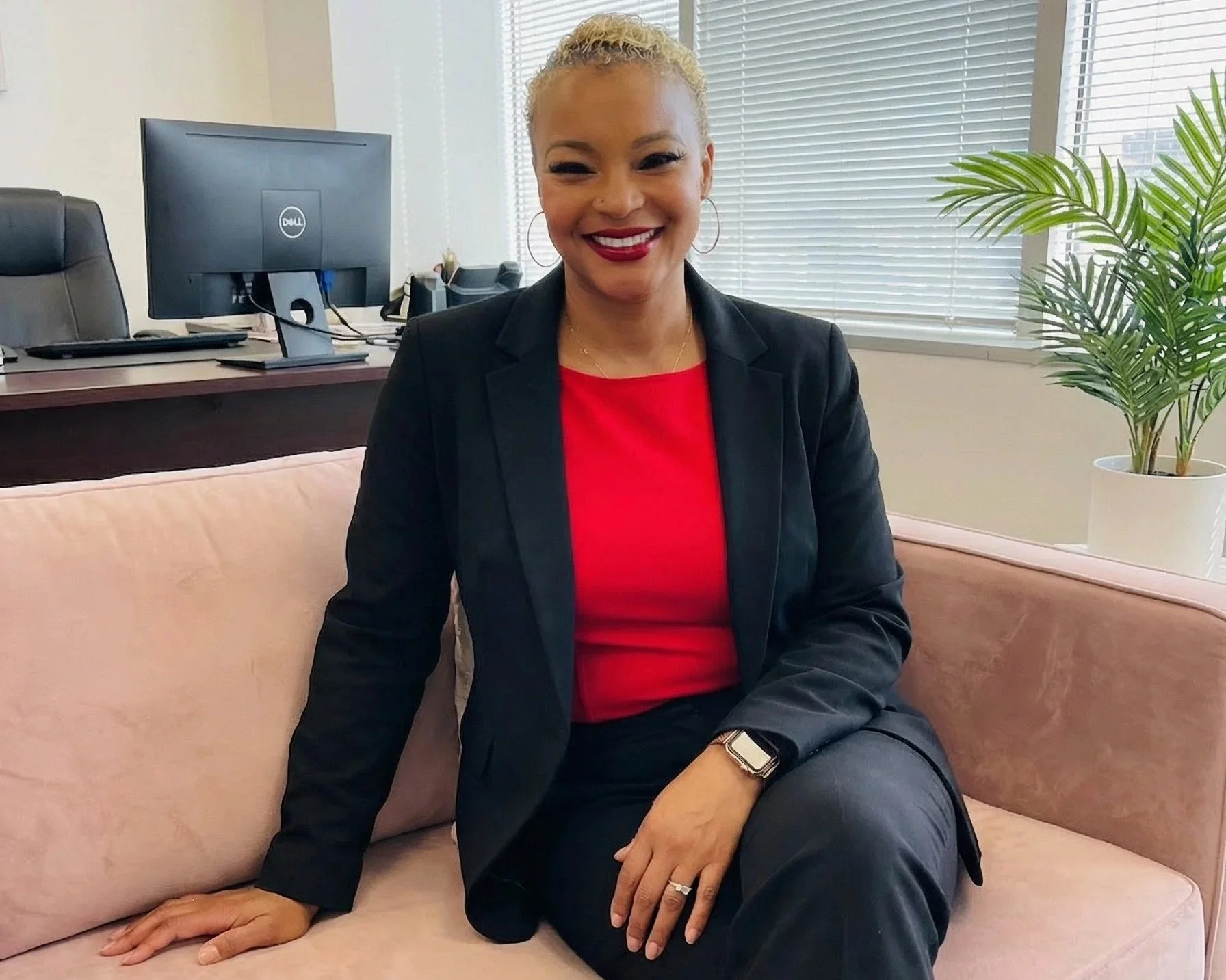 A woman with blonde curly hair smiling, dressed in a black blazer and red top, sitting on a pink sofa in an office, with a computer and a plant visible in the background.