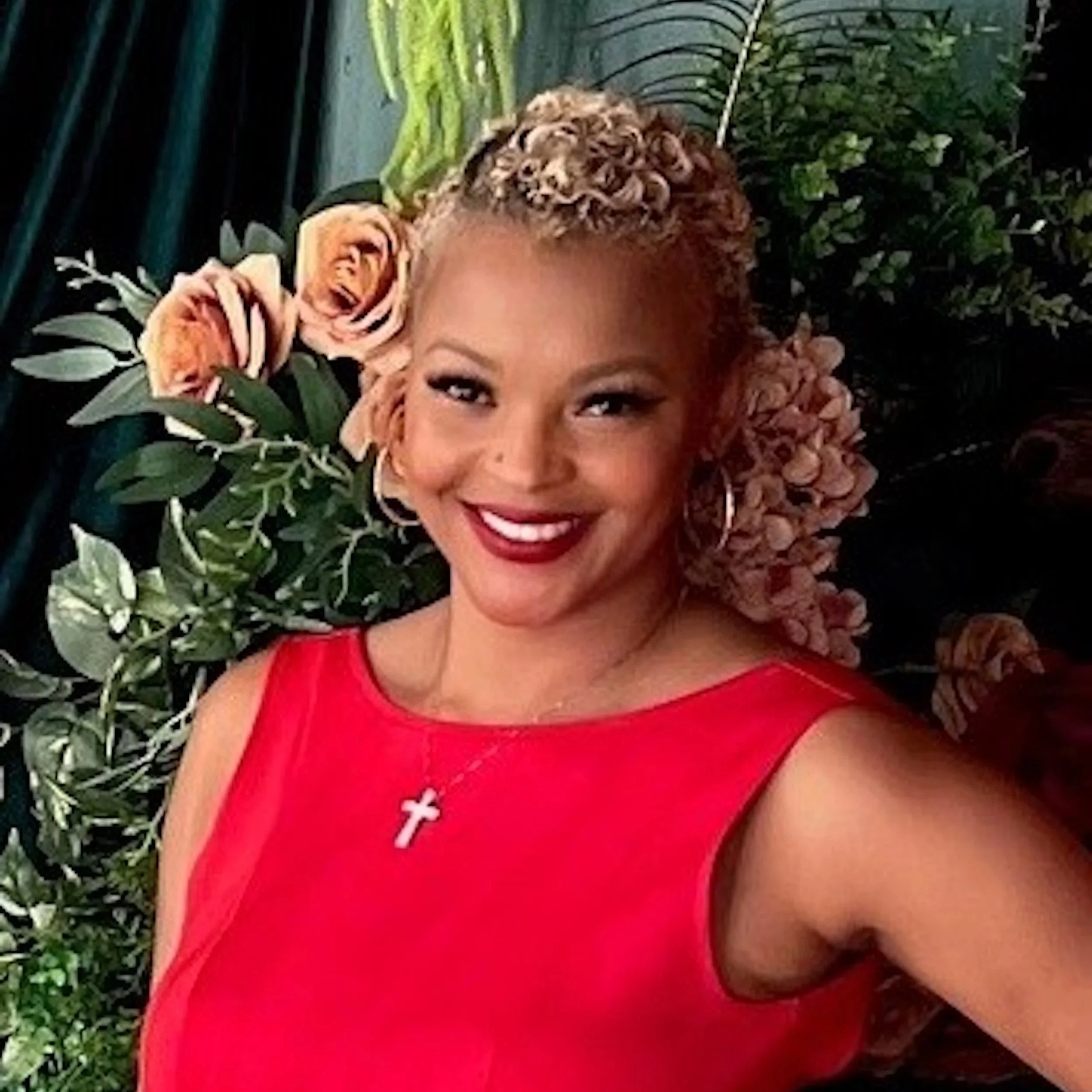 A woman with light skin and curly hair wearing a red top and a necklace with a cross pendant, smiling, surrounded by green plants and flowers.
