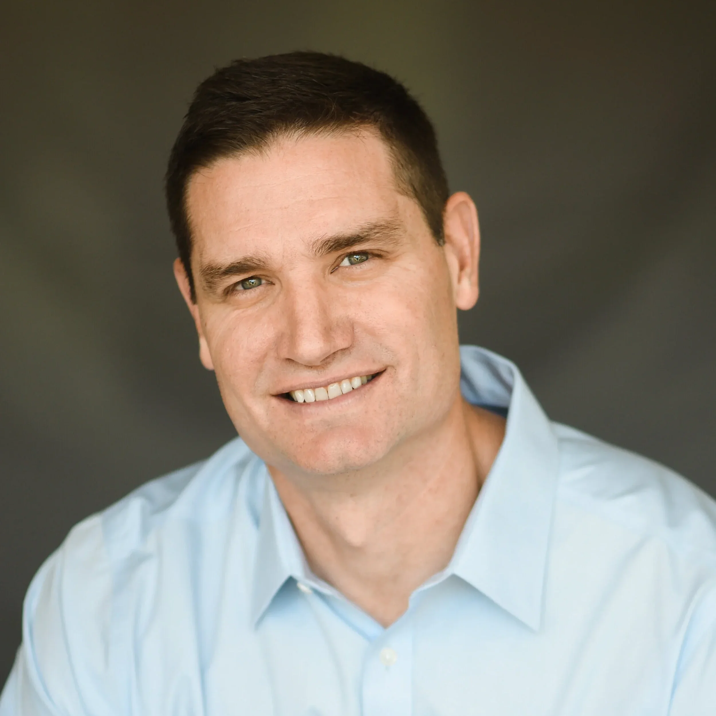 Close-up of a smiling man with short brown hair wearing a light blue shirt.