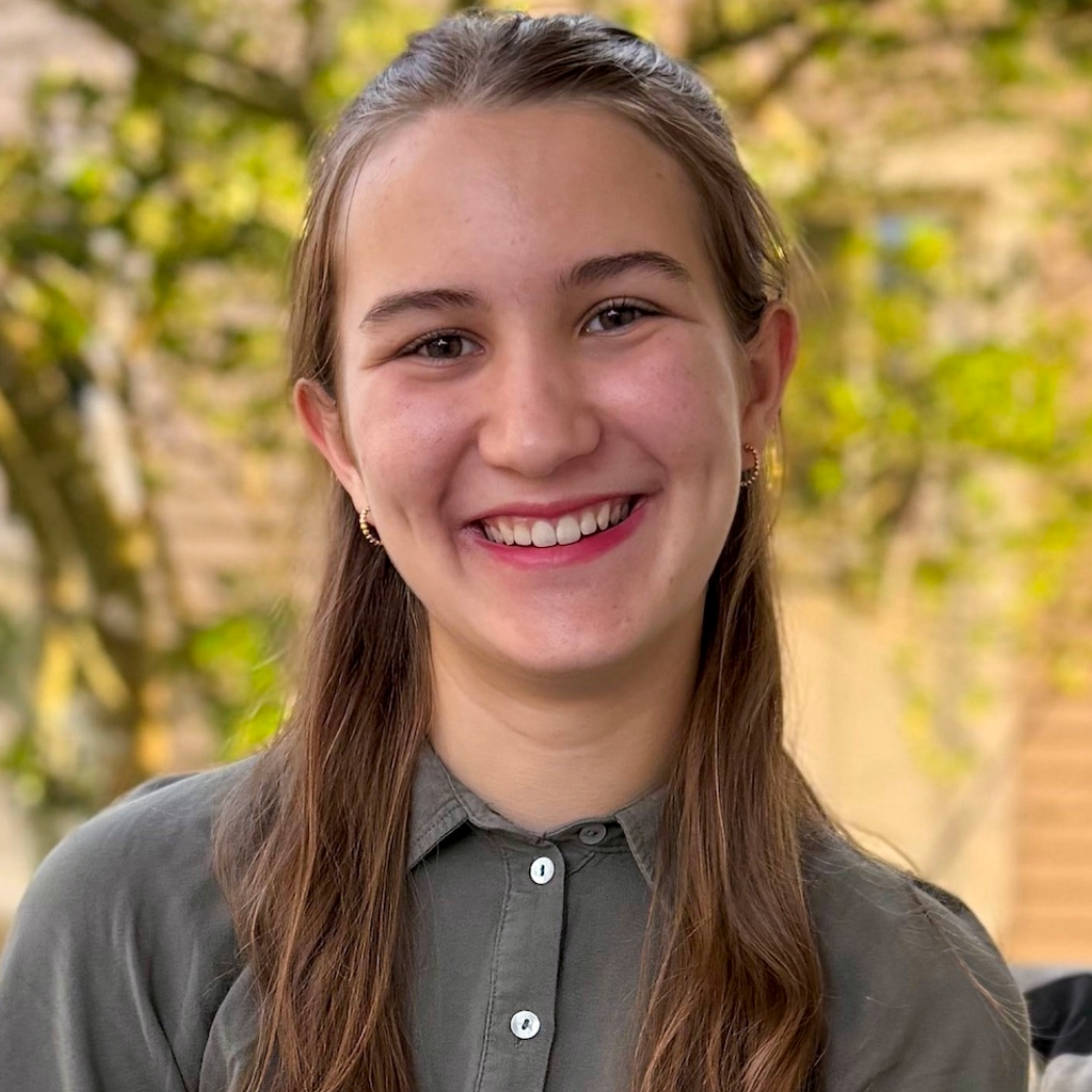 Young woman with long brown hair, wearing a gray button-up shirt, smiling outdoors with blurred trees and sunlight in the background.