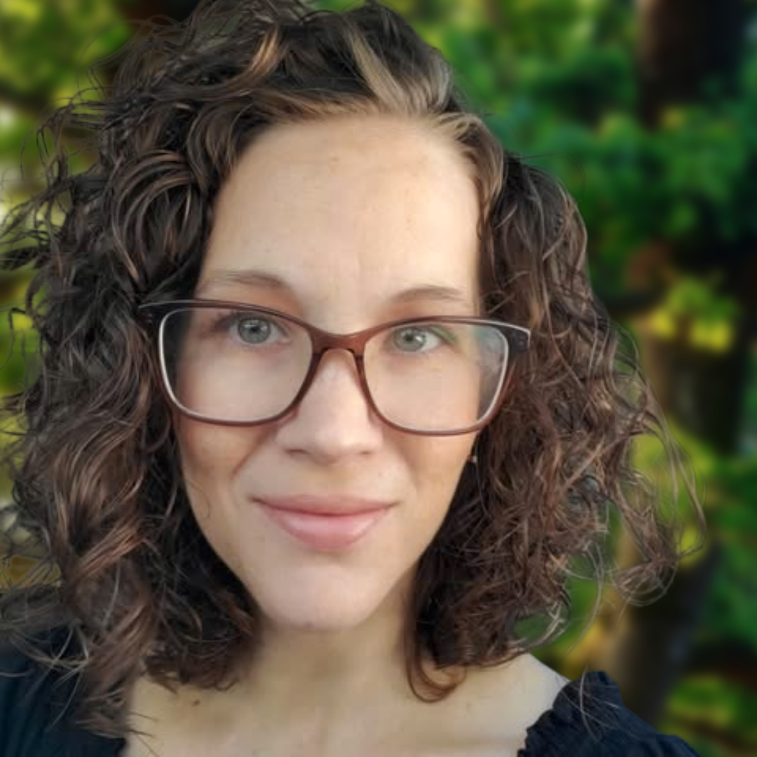 A woman with curly brown hair and glasses smiling outdoors, with green blurred foliage in the background.