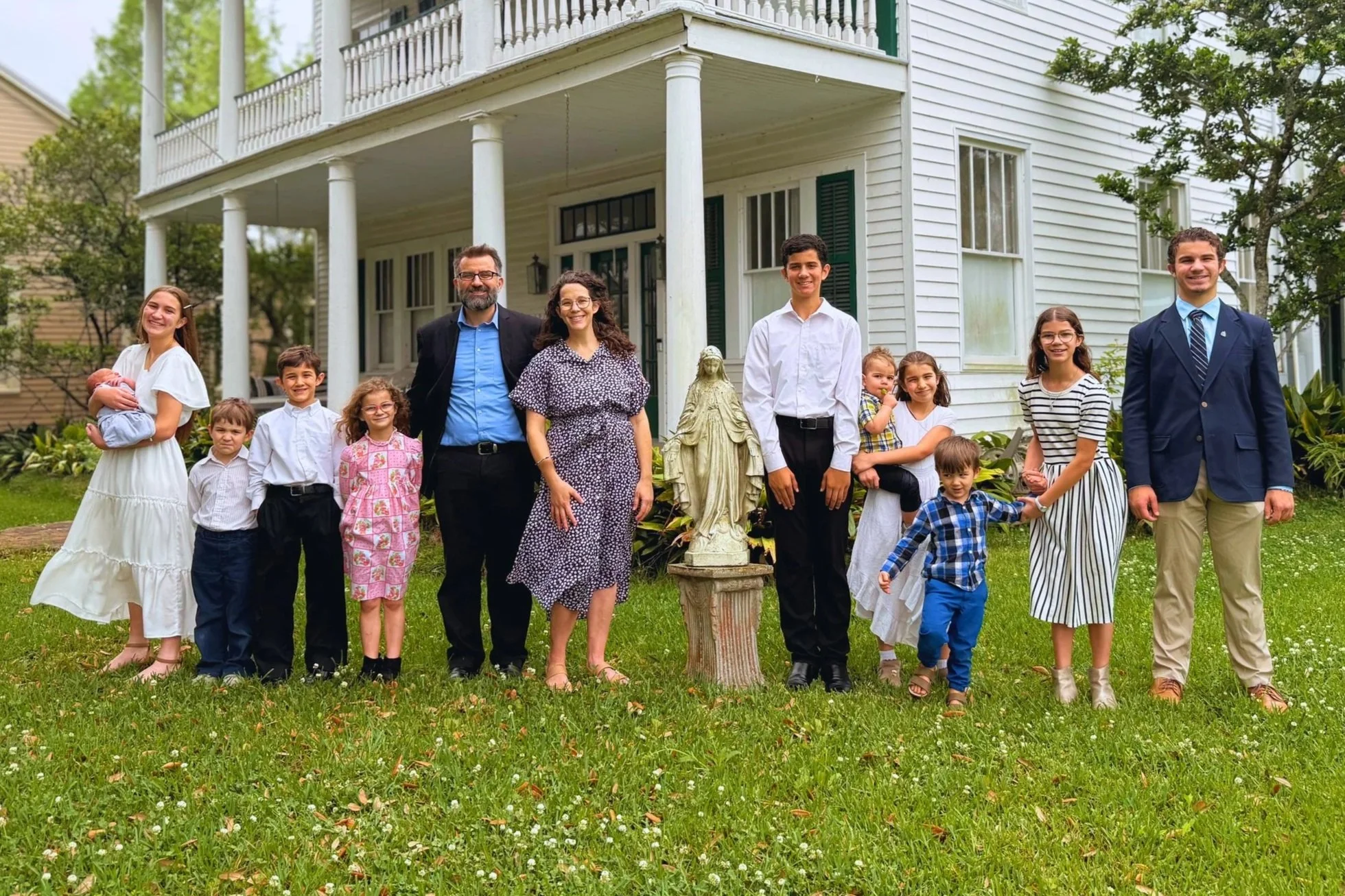 A large family group standing on the lawn in front of a white house with columns, smiling and posing for a photo. The group includes adults and children, some holding young children, with greenery and trees around.