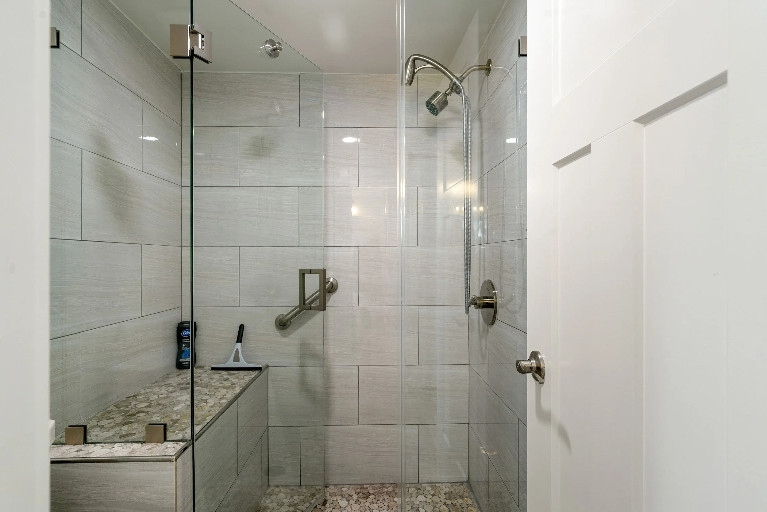 View of a modern shower with clear glass door, tiled walls, a showerhead, a grab bar, and cleaning tools inside the shower area.