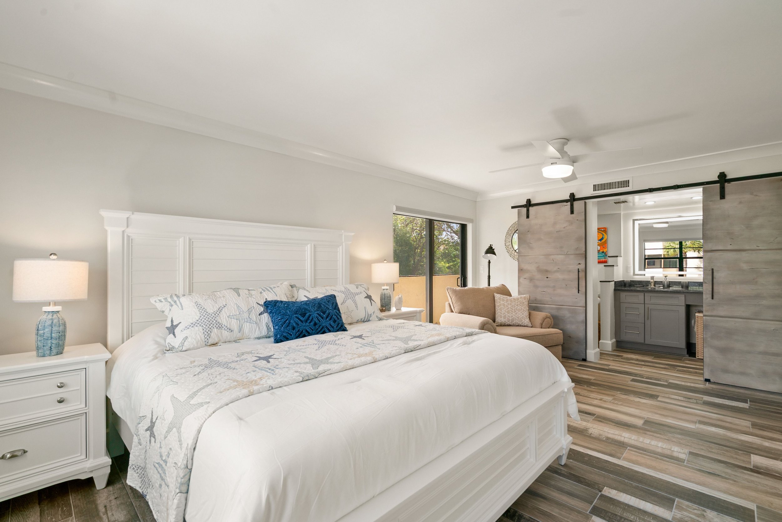 A bedroom with a white bed frame and starfish-patterned bedding, a nightstand with a lamp, beige armchair, sliding barn door, and a ceiling fan, with a view of the living area and kitchen.