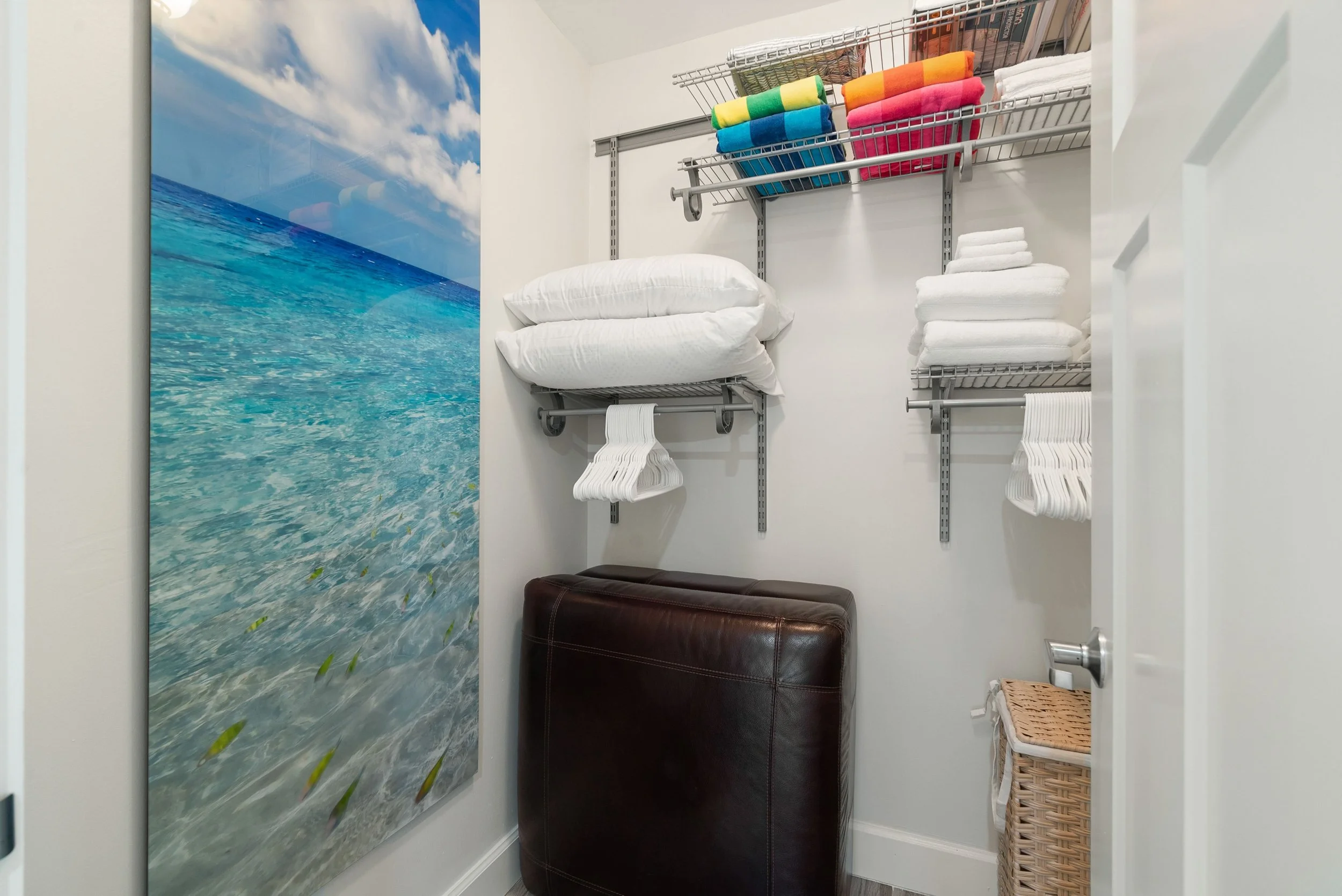 A closet with a beach mural on the wall, organized shelving with folded towels, a pillow on a metal shelf, a small brown ottoman, and a wicker basket.