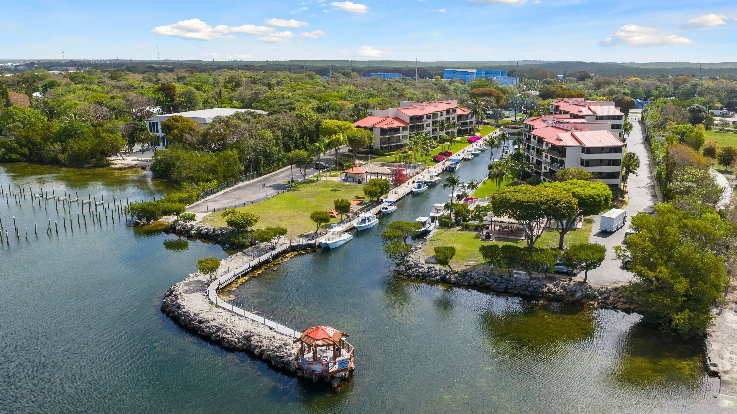 Aerial view of a waterfront residential area with multi-story buildings, boats docked along a canal, surrounded by lush green trees, a small gazebo on a pier, and a parking lot in the background under a partly cloudy sky.