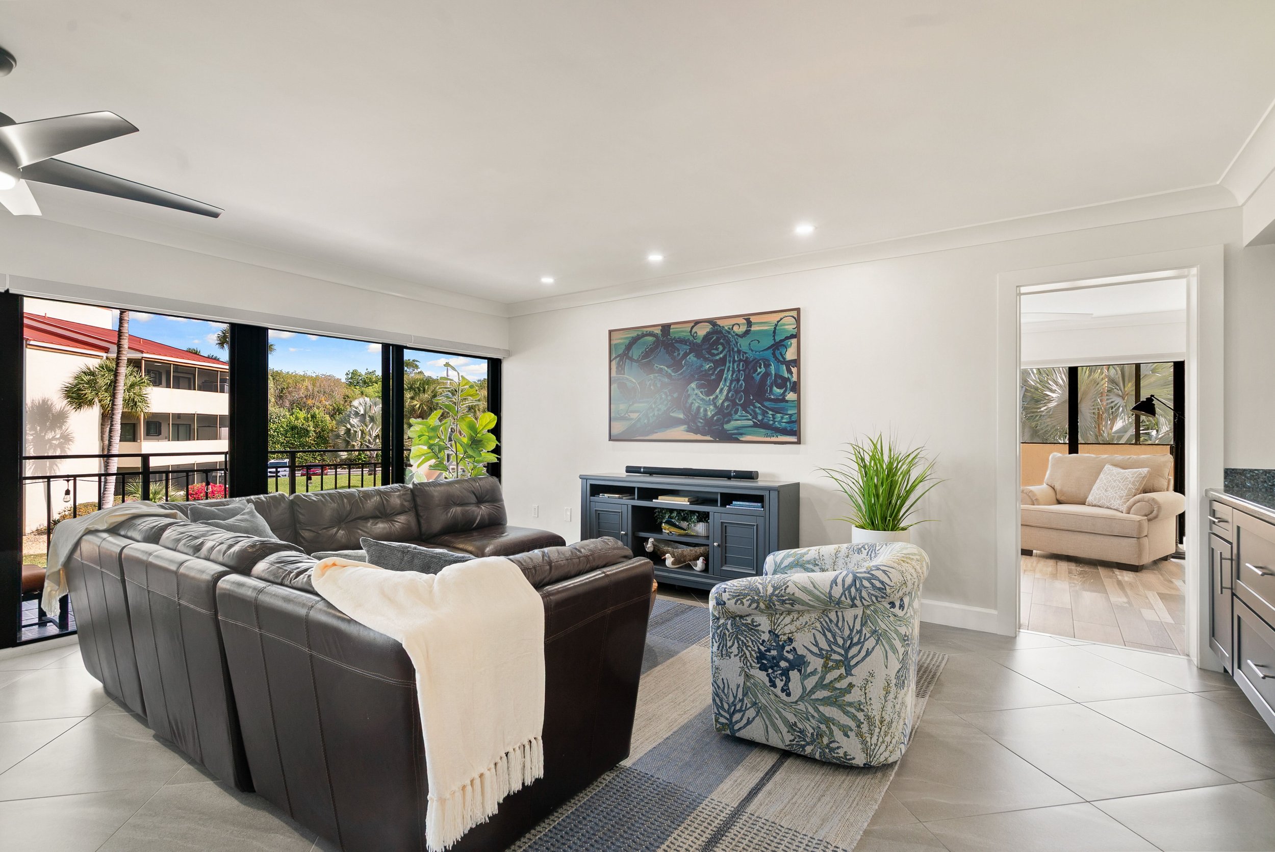 Living room with large windows, tan leather sofa, patterned armchair, black media stand with a soundbar, wall art of an octopus, potted plants, and a view of outdoor trees and buildings