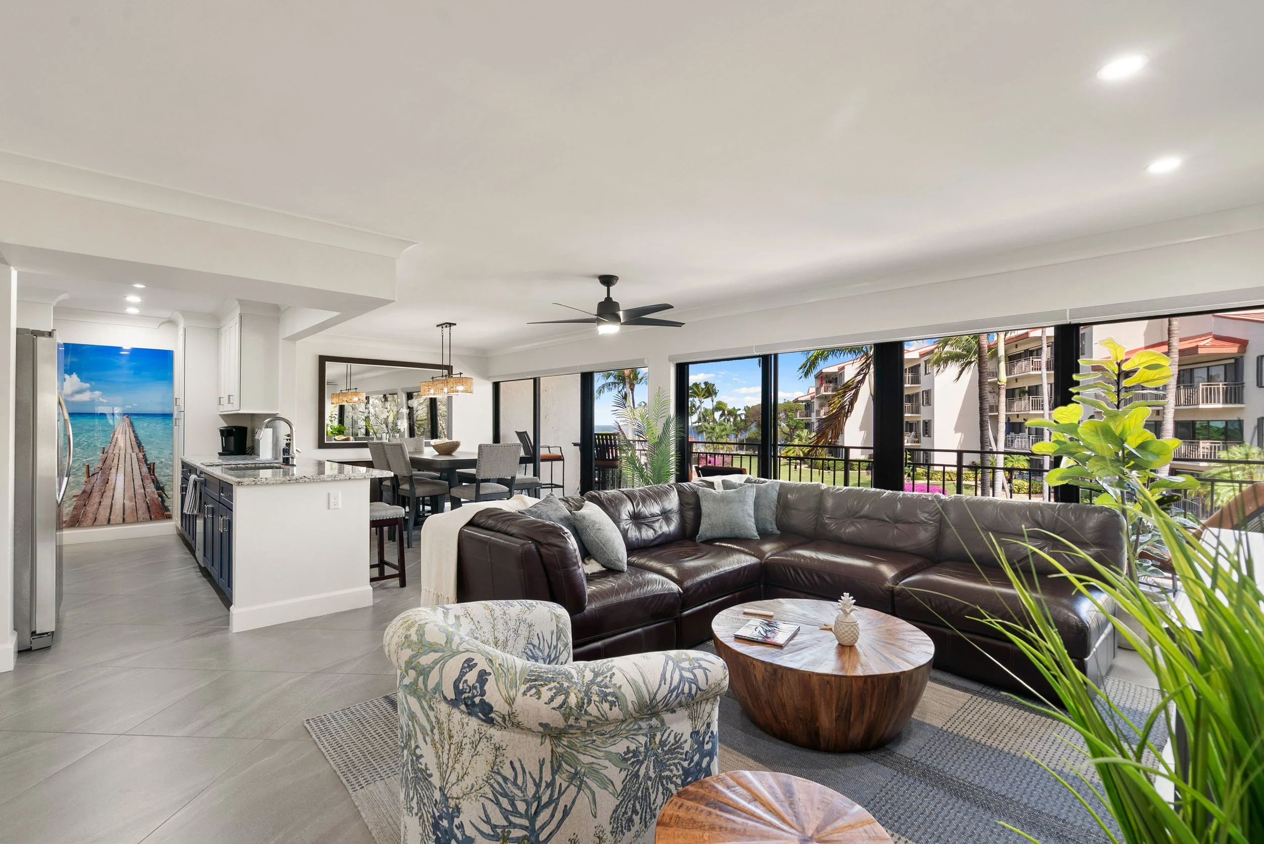 Open-concept living room and kitchen with large windows showing a view of buildings, palm trees, and a blue sky.