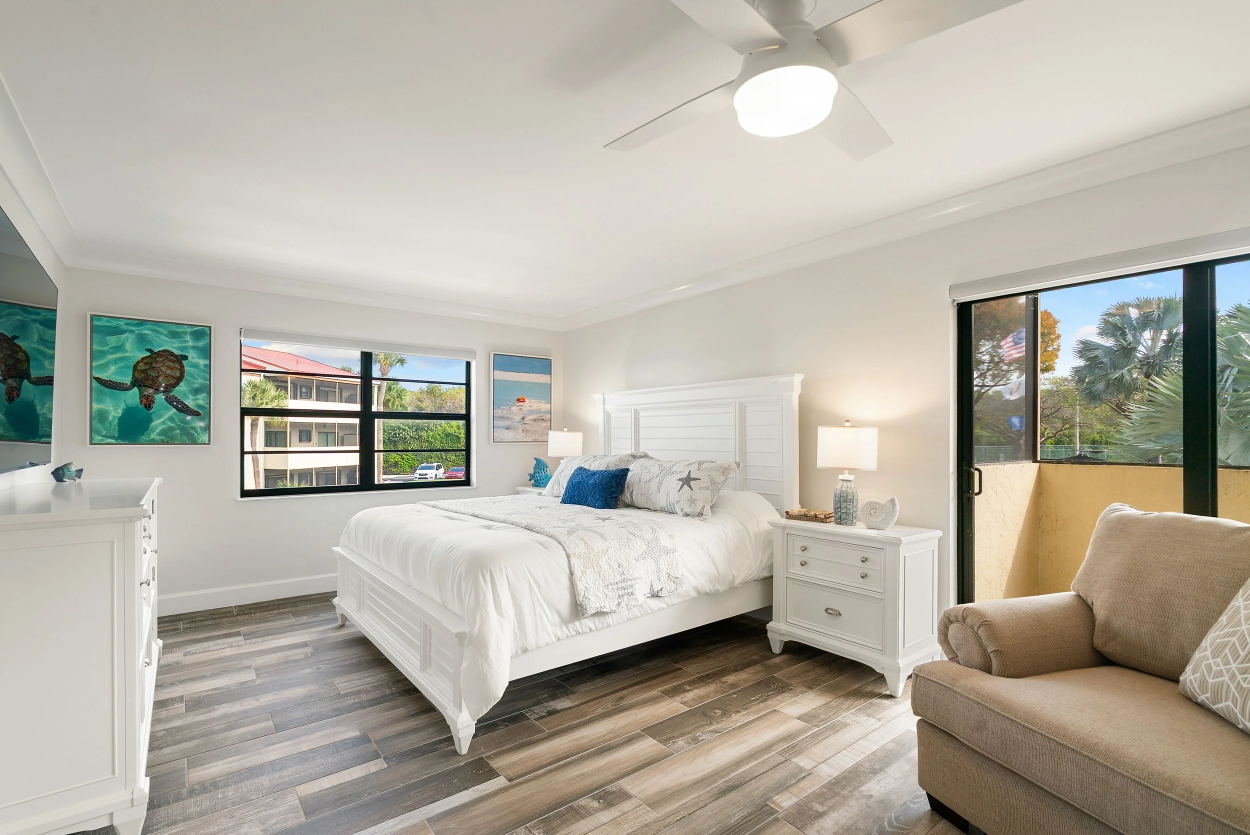 A bright bedroom with white walls and furniture, including a bed with white bedding and starfish print pillows, side tables with lamps, large windows showing trees, a palm tree outside, and artwork of turtles on the wall.