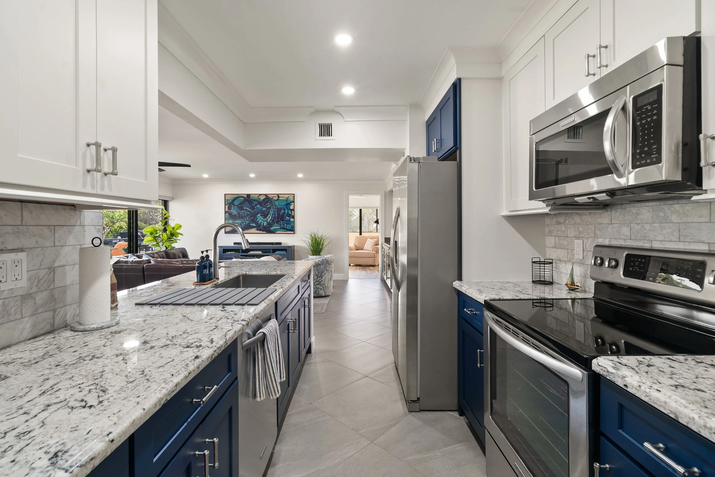 Modern kitchen with granite countertops, white cabinets, blue lower cabinets, stainless steel appliances including a microwave and oven, with an open view into a living room area that has a painting of an octopus and seating.