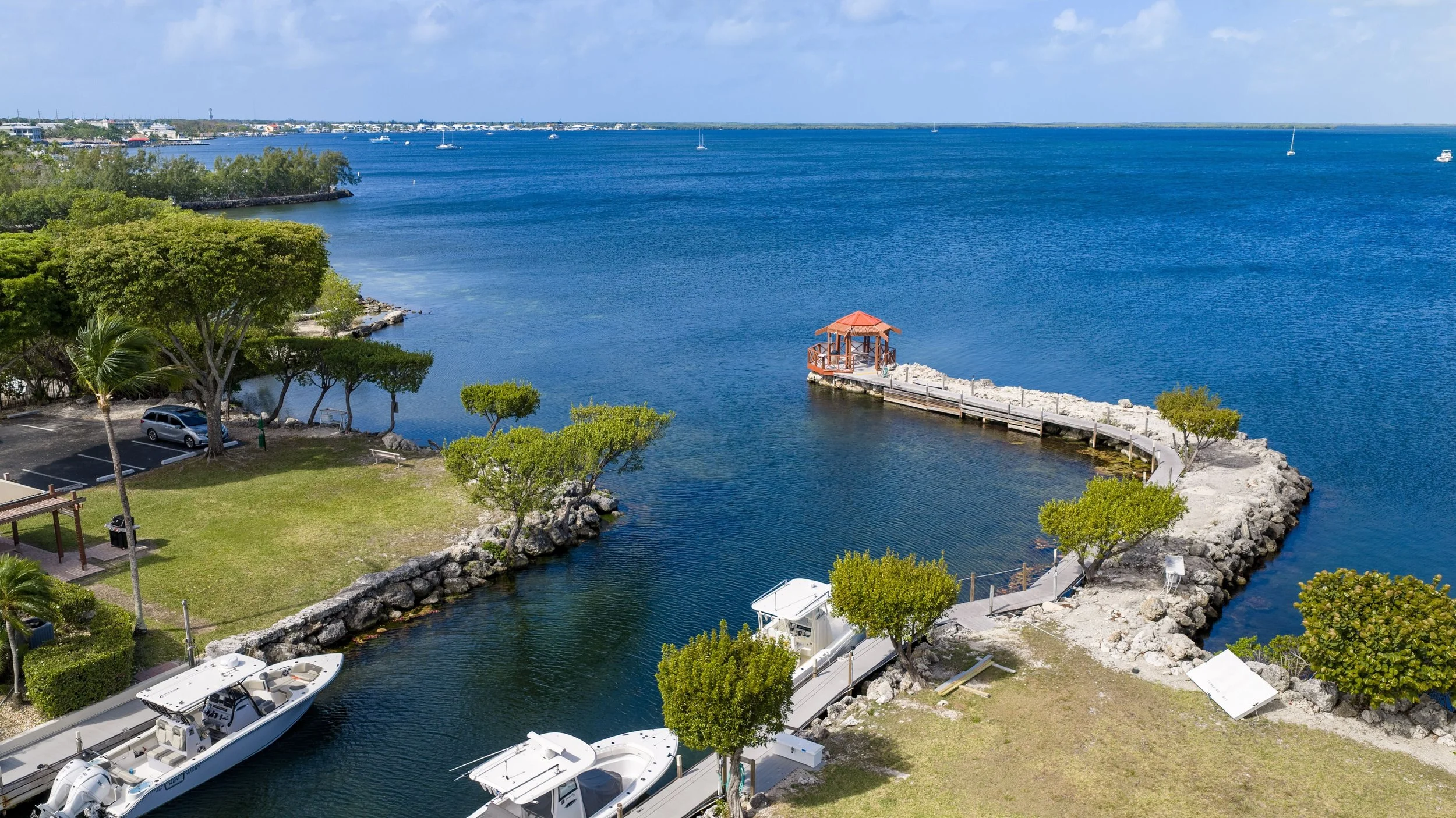 A marina with boats docked along a waterway, a pier with a small gazebo, green trees, and a cityscape across the water under a partly cloudy sky.