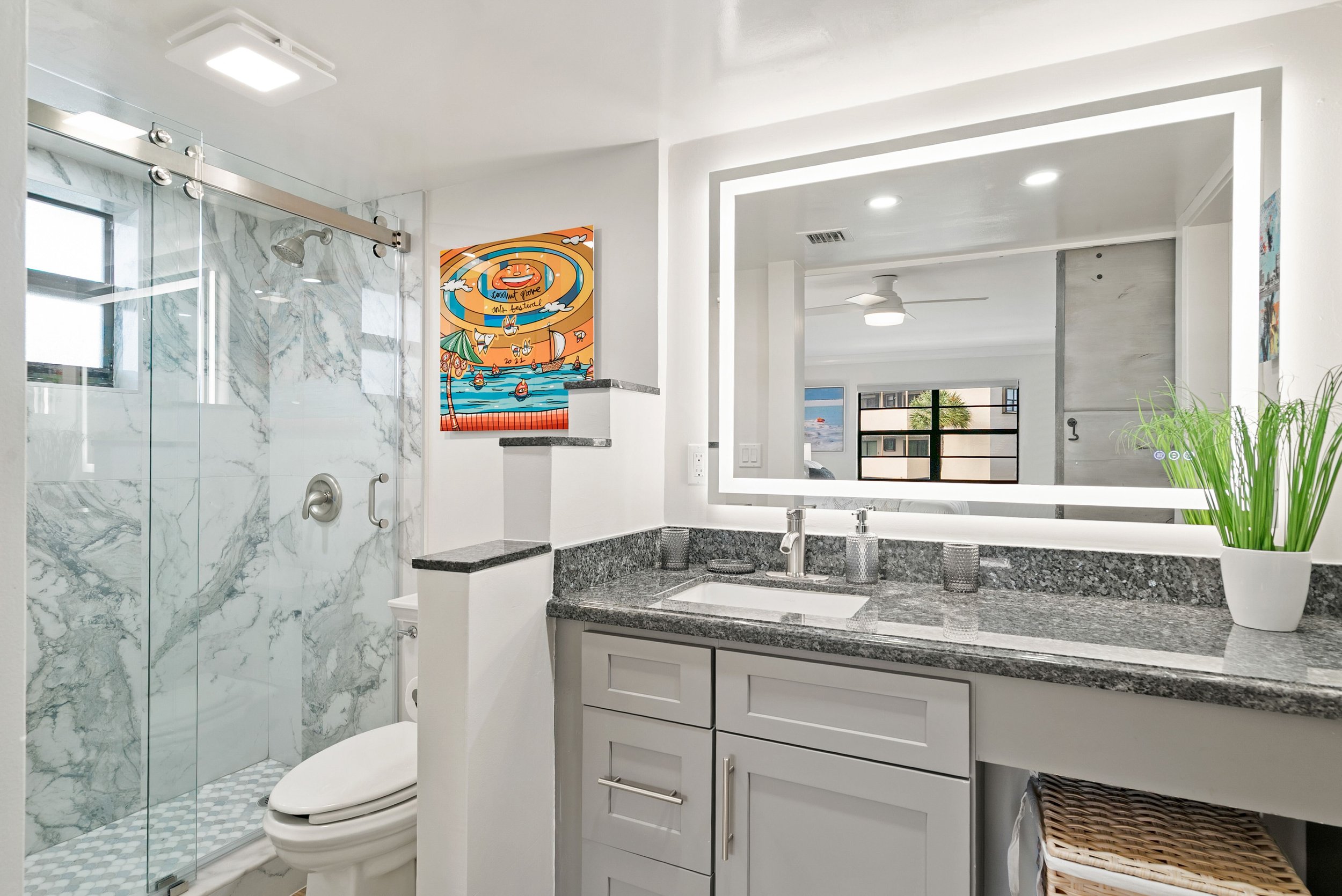 A modern bathroom with a marble shower enclosure, a vanity with a granite countertop, and a large illuminated mirror. There is a colorful abstract painting on the wall, a window, and a potted plant on the right side.