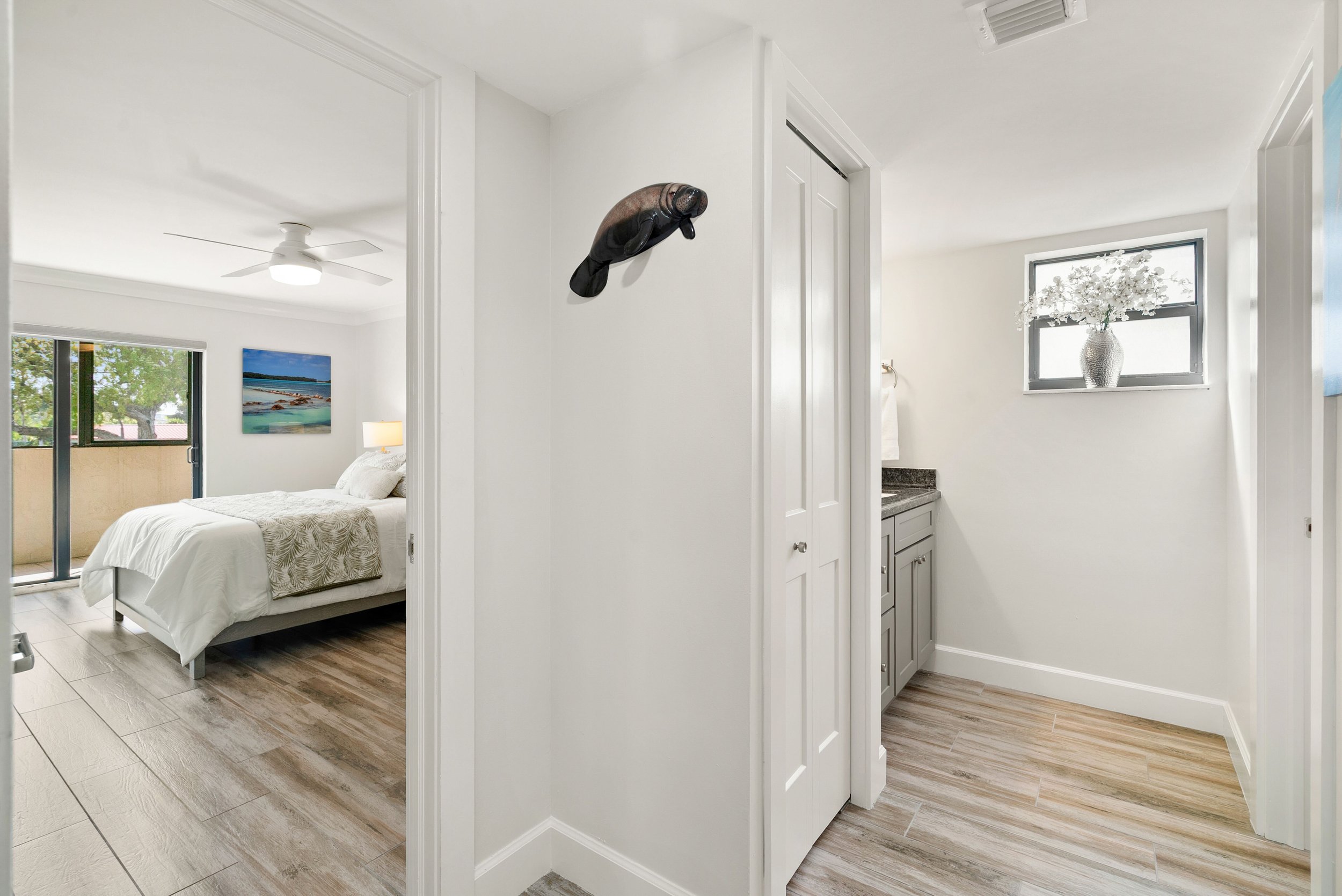 The image shows a bedroom with a bed, a window, a ceiling fan, and a wall hanging of a seal, with a partial view of a bathroom with a window and a floral vase.