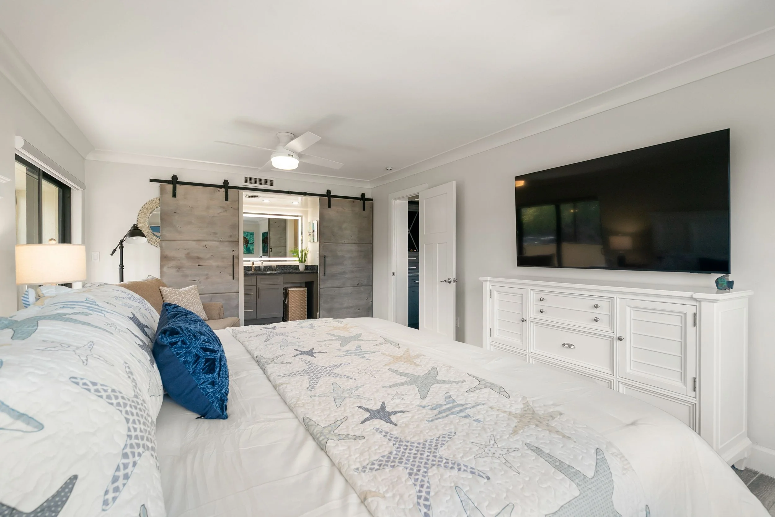 A bedroom featuring a large bed with starfish-patterned bedding and pillows, a flat-screen TV mounted on the wall, a sliding barn door leading into another room, a white dresser, and a ceiling fan.