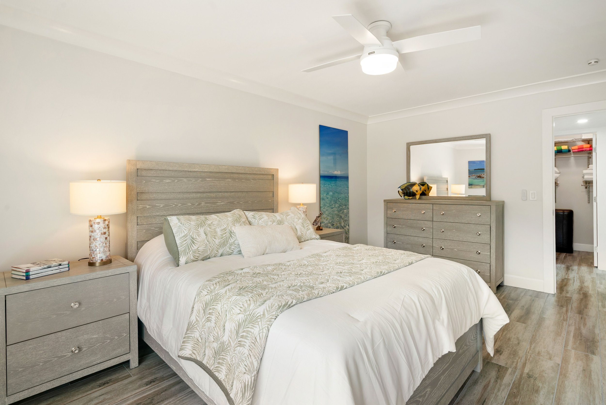 A bedroom with a large bed with light-colored, patterned bedding, a gray wood headboard, matching gray wood nightstand with a lamp and magazines, a matching gray wood dresser with a large mirror and decorative fish, a wall-mounted art piece of a beac
