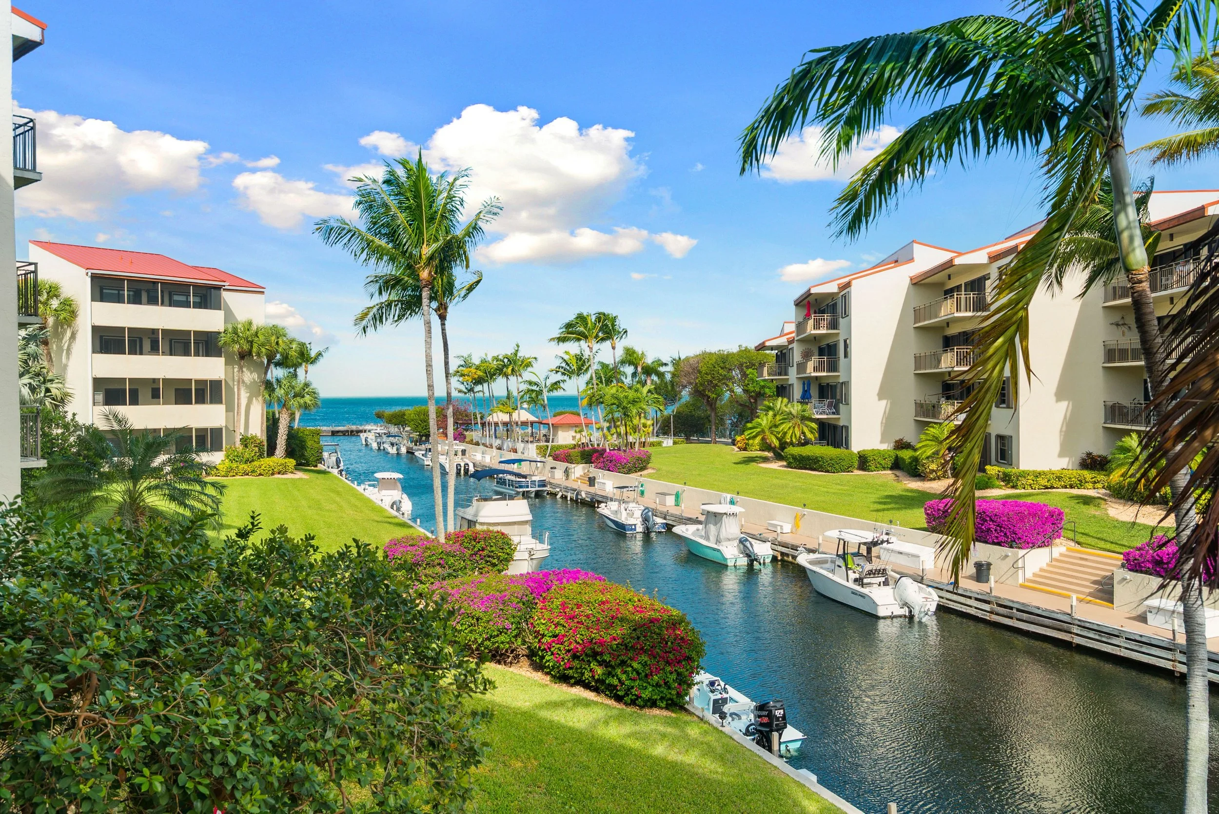 A view of a tropical resort with white buildings, palm trees, a canal with boats, colorful flowers, and the ocean in the background under a partly cloudy sky.