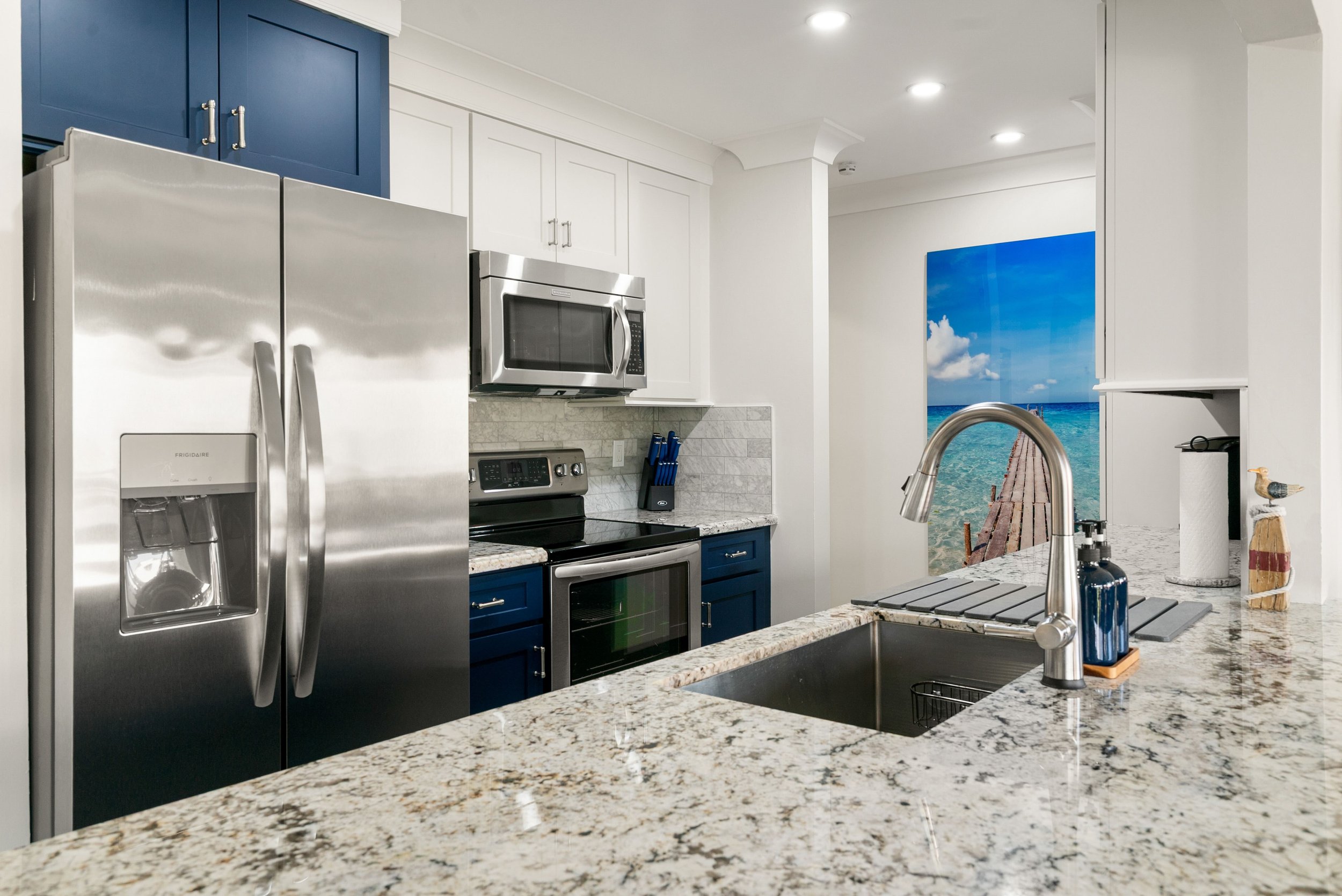 Kitchen with granite countertops, stainless steel refrigerator, microwave, stove, blue and white cabinets, and a wall mural of a beach pier leading to the ocean.
