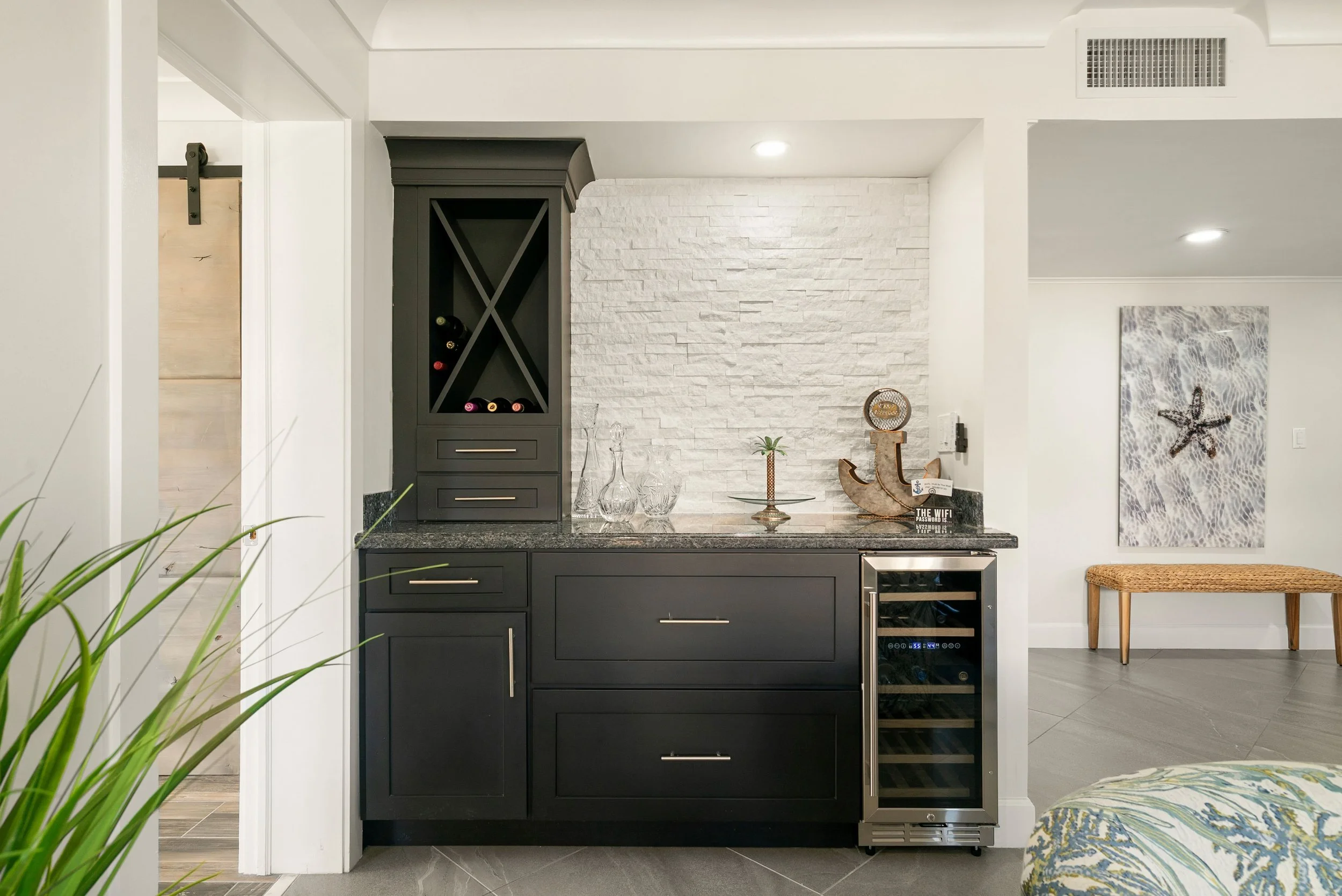 A kitchen with dark grey cabinets, a white textured brick wall, a wine rack with bottles, decorative glass vases, and a small wine fridge underneath a granite countertop. There is a piece of artwork with a starfish on the right and a woven bench on t