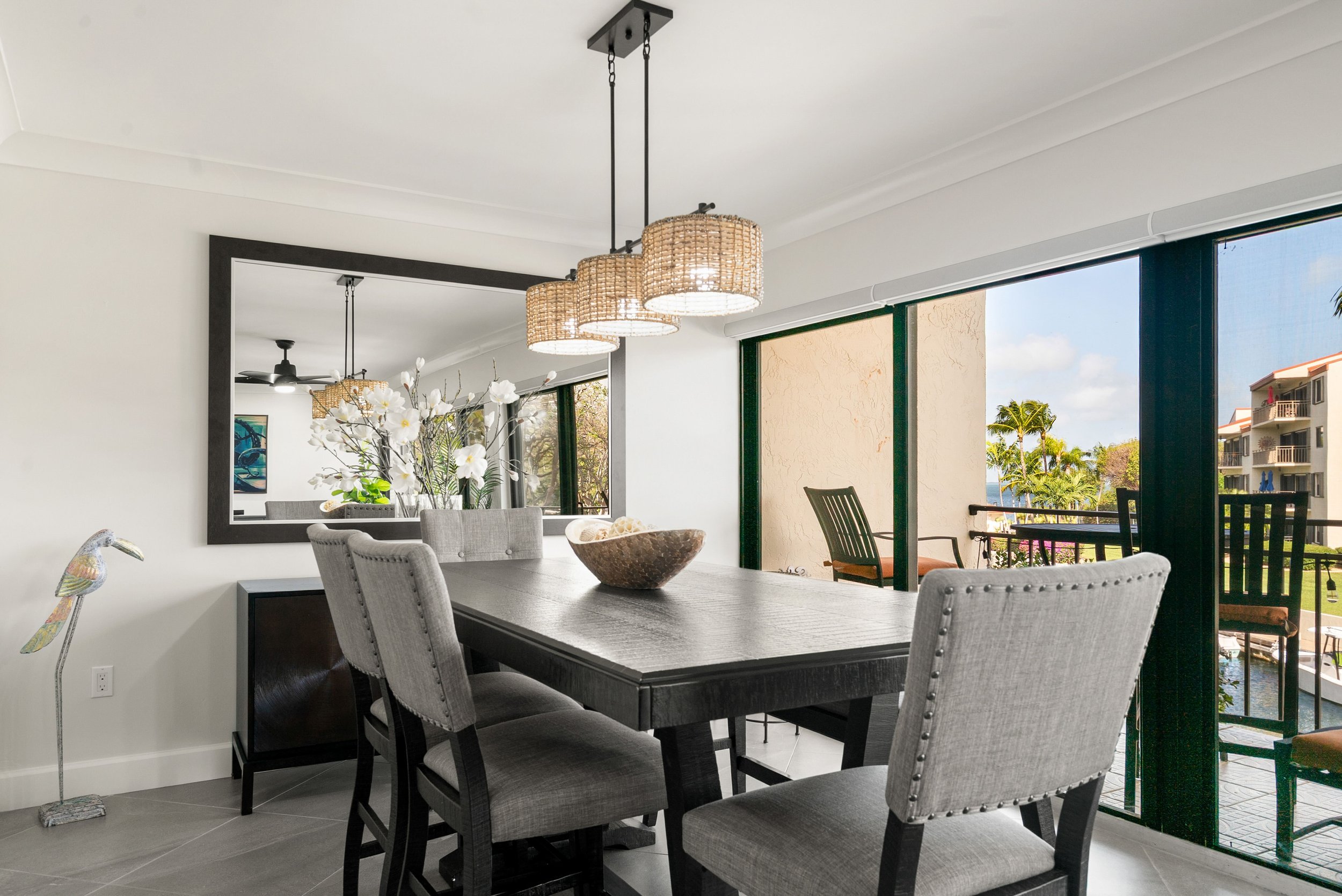 A dining room with a dark wooden table, six gray upholstered chairs, a large mirror on the wall, a woven pendant light hanging from the ceiling, a bowl of decorative items on the table, and a sliding glass door leading to a balcony with outdoor furni