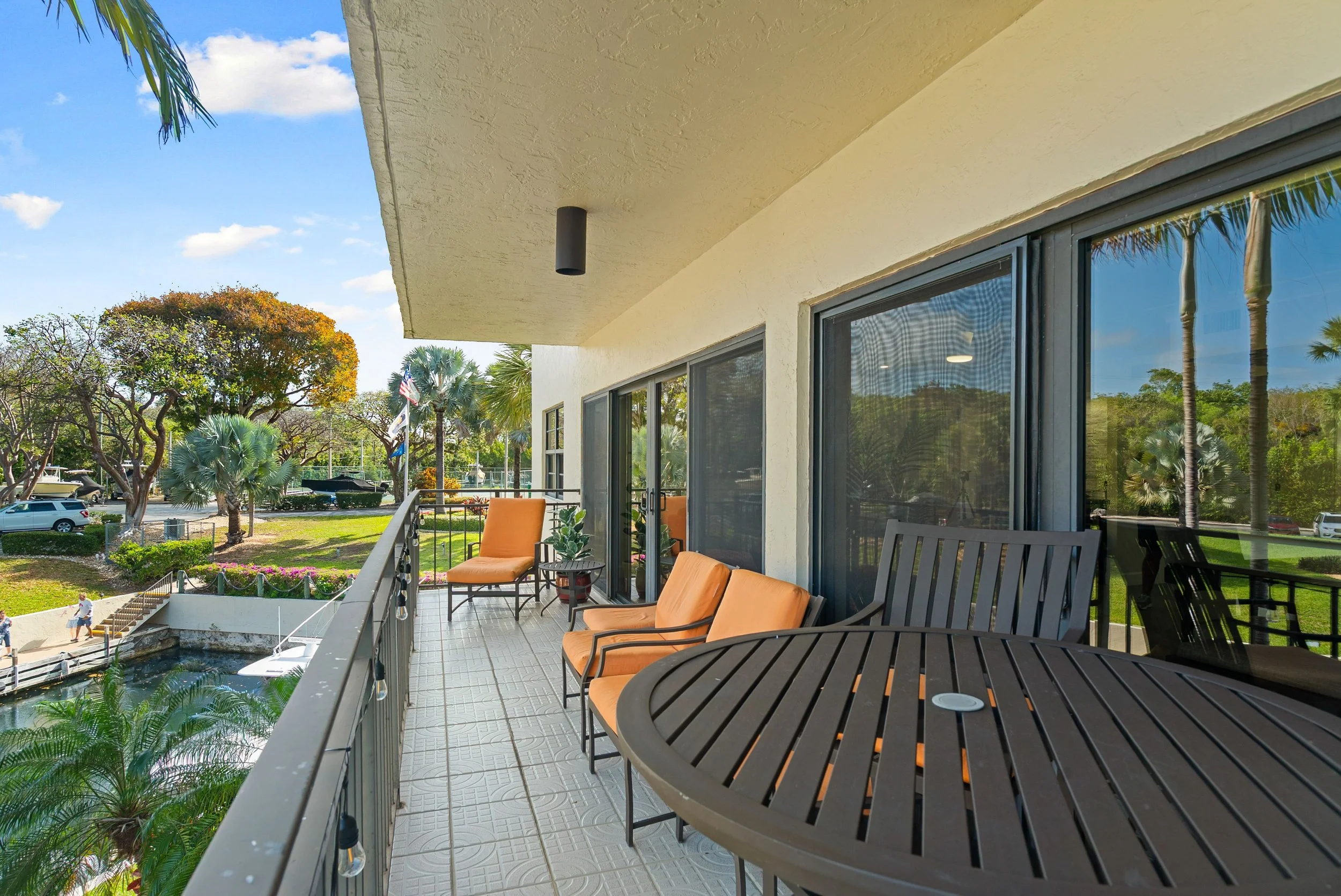 Balcony with black metal chairs and orange cushions, overlooking a garden with trees, parking lot, and tennis courts under a partly cloudy sky.