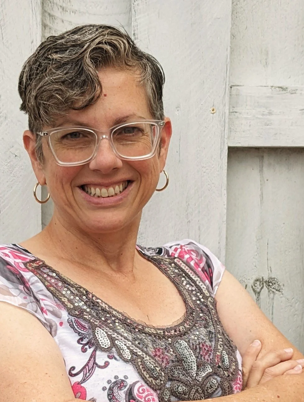 Smiling woman with short gray hair, glasses, and hoop earrings, wearing a patterned top, standing against a light wood background.