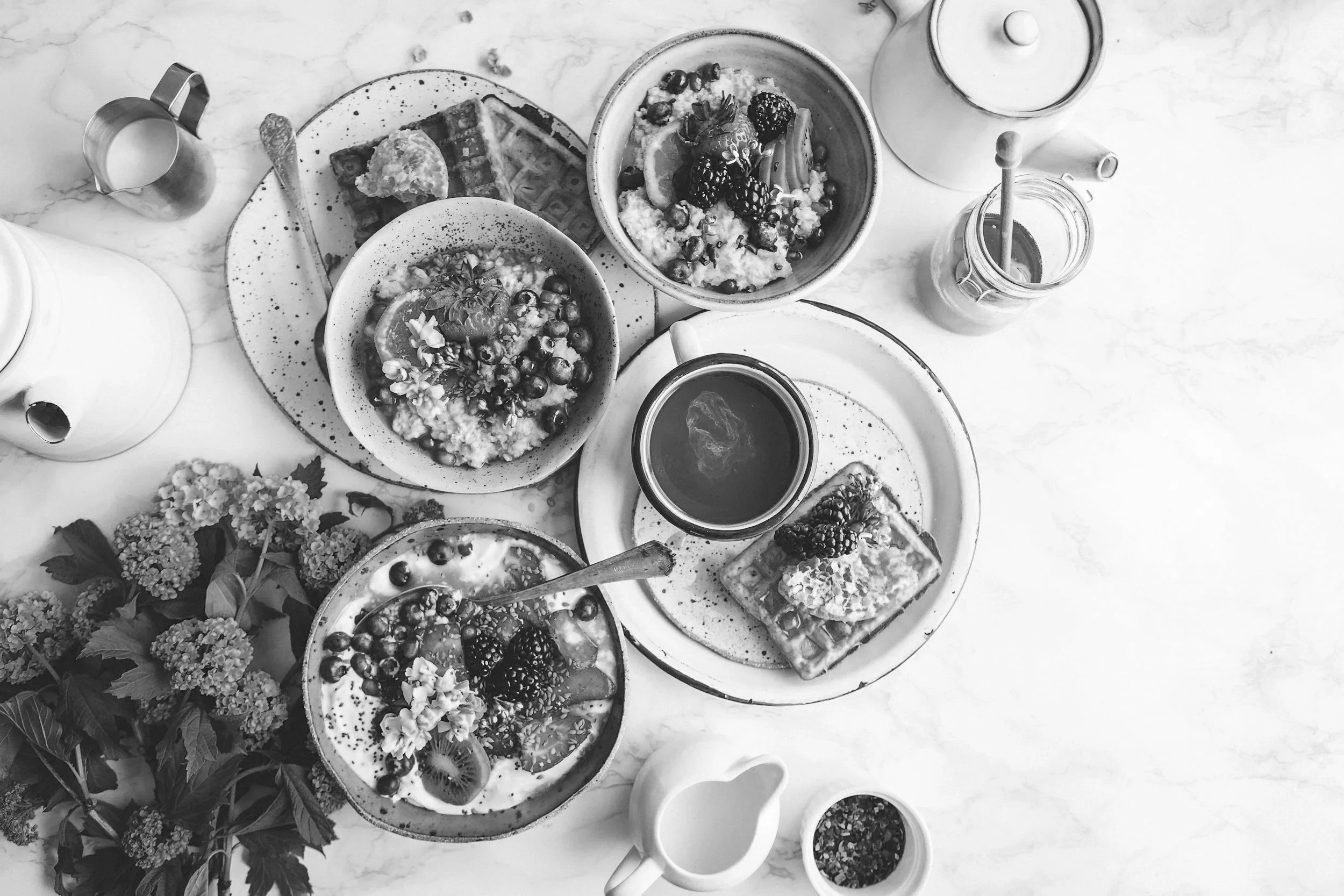 Table de petit-déjeuner avec plusieurs bols de céréales, fruits, gaufres, une tasse, une théière, un pot à lait, un vase avec des fleurs, et plusieurs autres accessoires de cuisine.