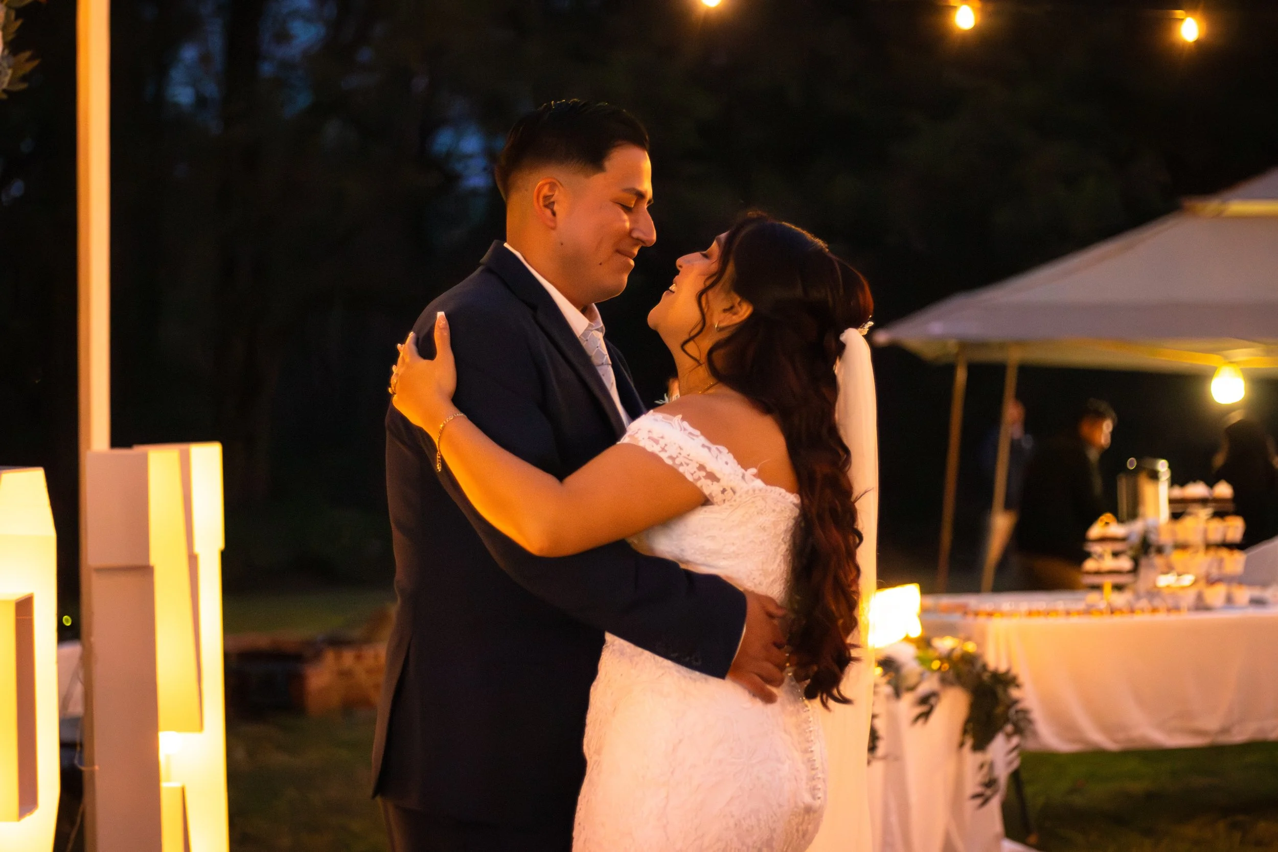 A bride and groom dancing closely at their wedding reception outdoors at night, with warm lighting and a dessert table in the background.