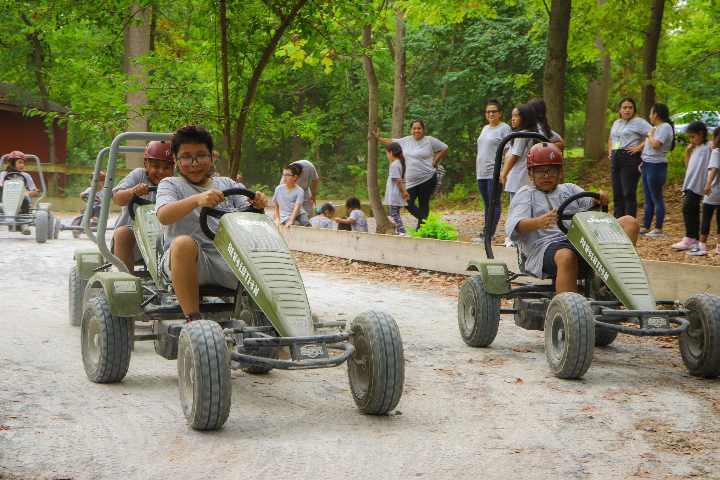 Children riding small, green go-karts on a dirt trail in a wooded outdoor area with adults and other children watching nearby.