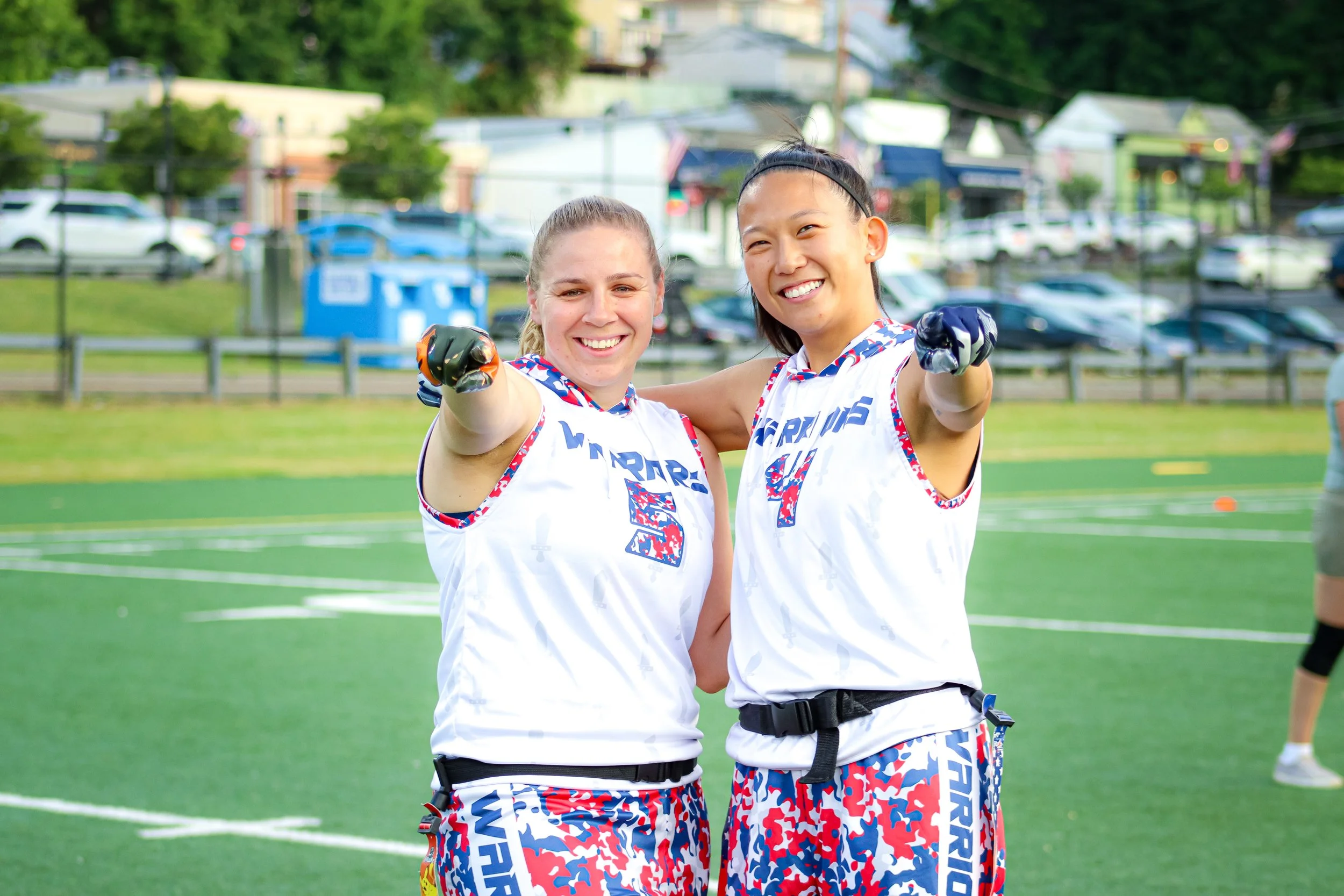 Two smiling female flag football players standing on a football field pointing forward.