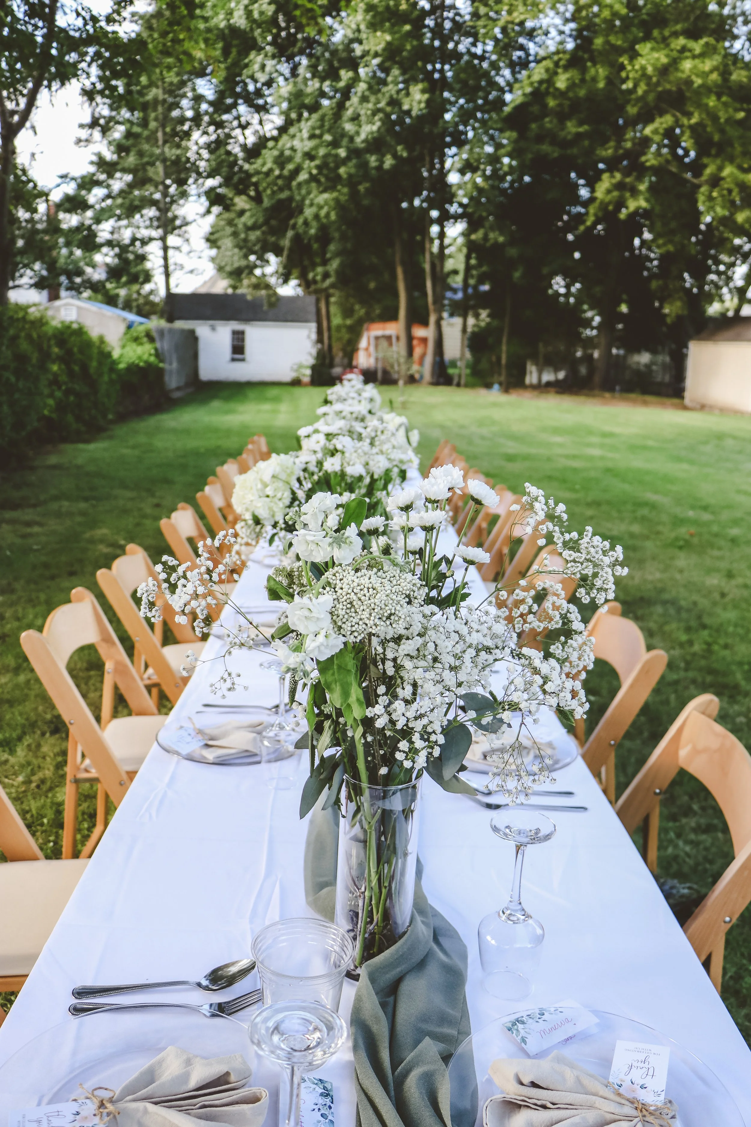 Outdoor long dining table decorated with white floral centerpieces and place settings, set in a grassy backyard with trees and buildings in the background.