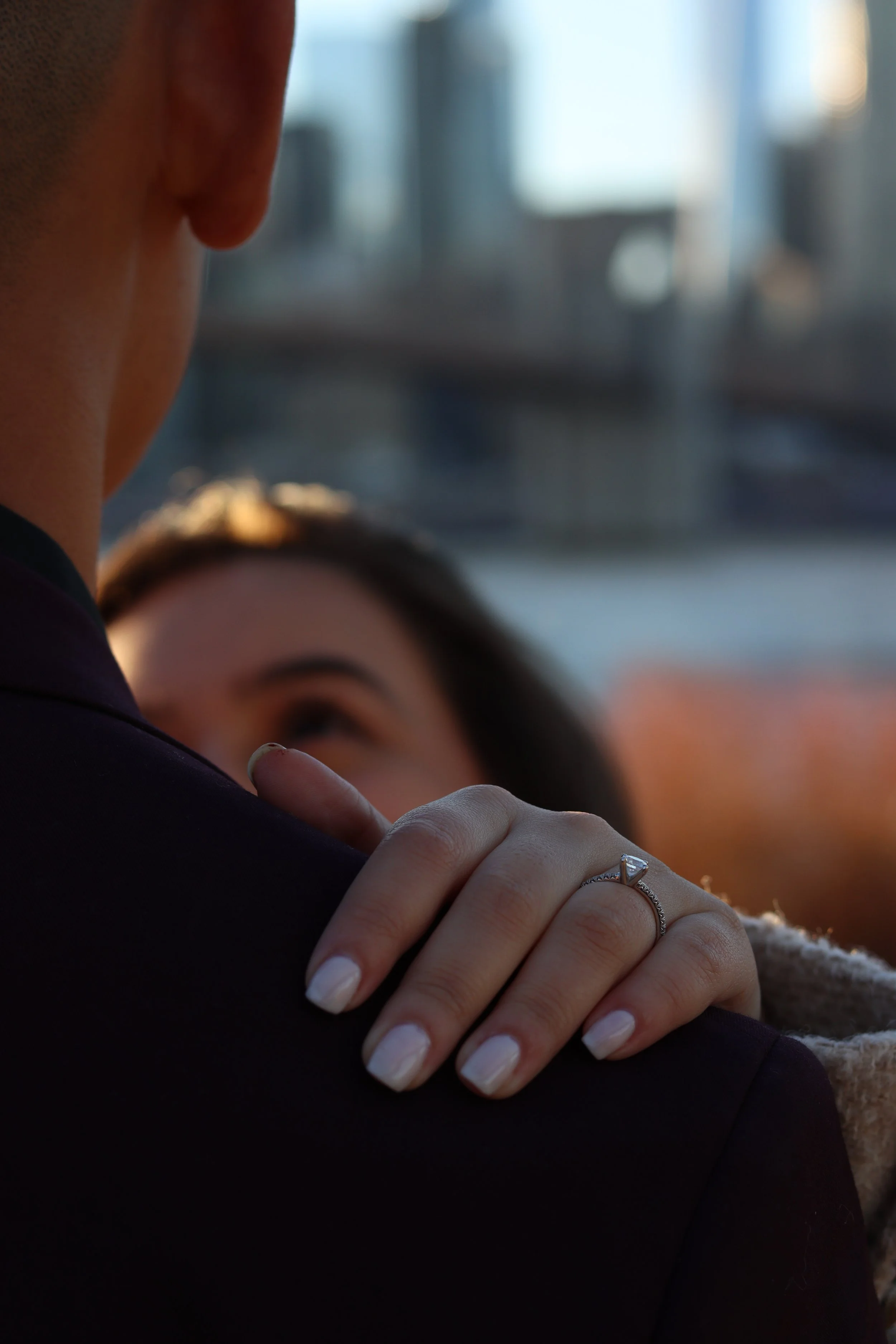 A woman with painted nails resting her hand on a man's shoulder, showing a diamond engagement ring, both embracing near a window with cityscape in the background.
