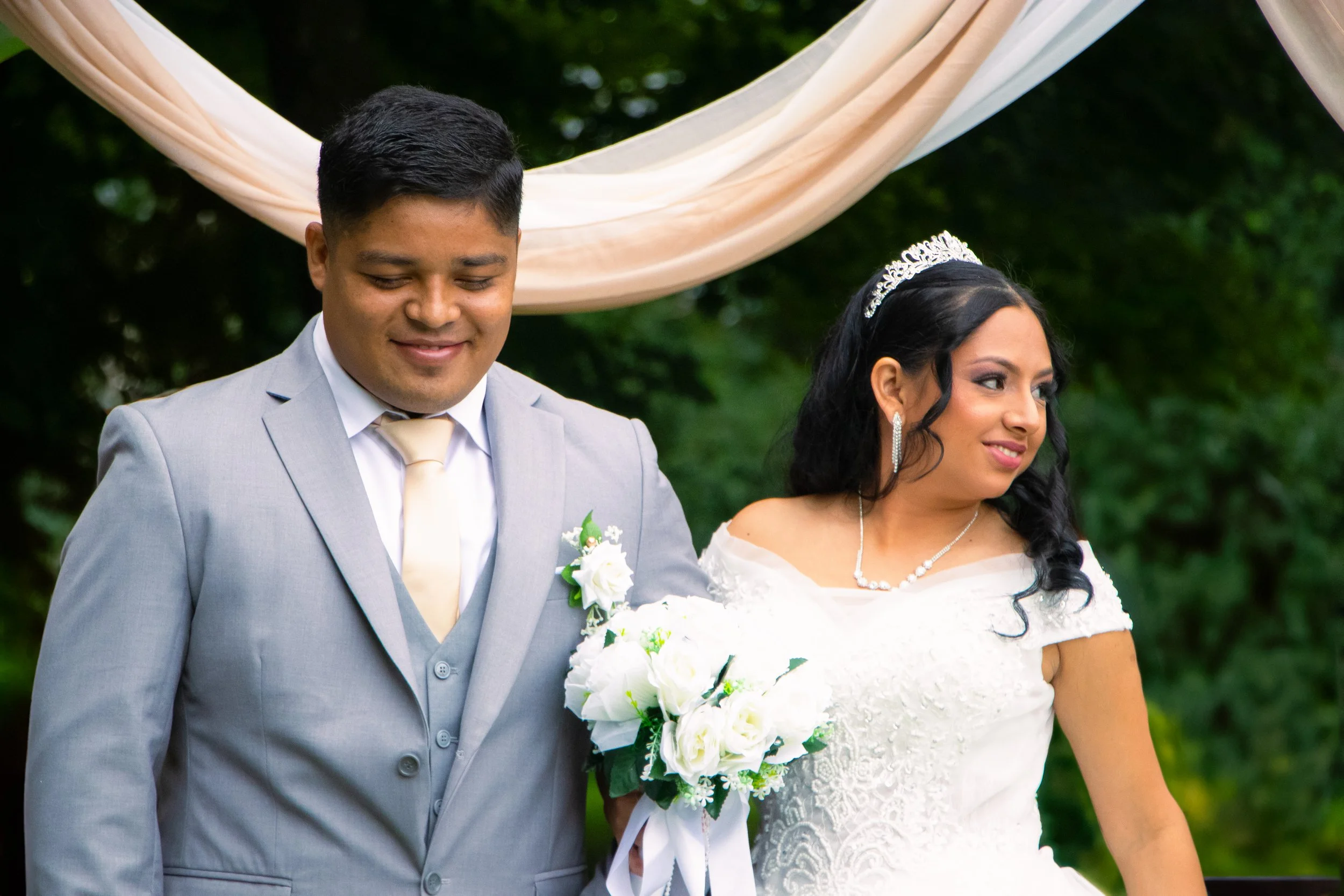 A bride and groom standing together outdoors during their wedding, with the bride holding a bouquet of white roses and wearing a white wedding gown and tiara, and the groom wearing a light gray suit with a white shirt and tan tie.