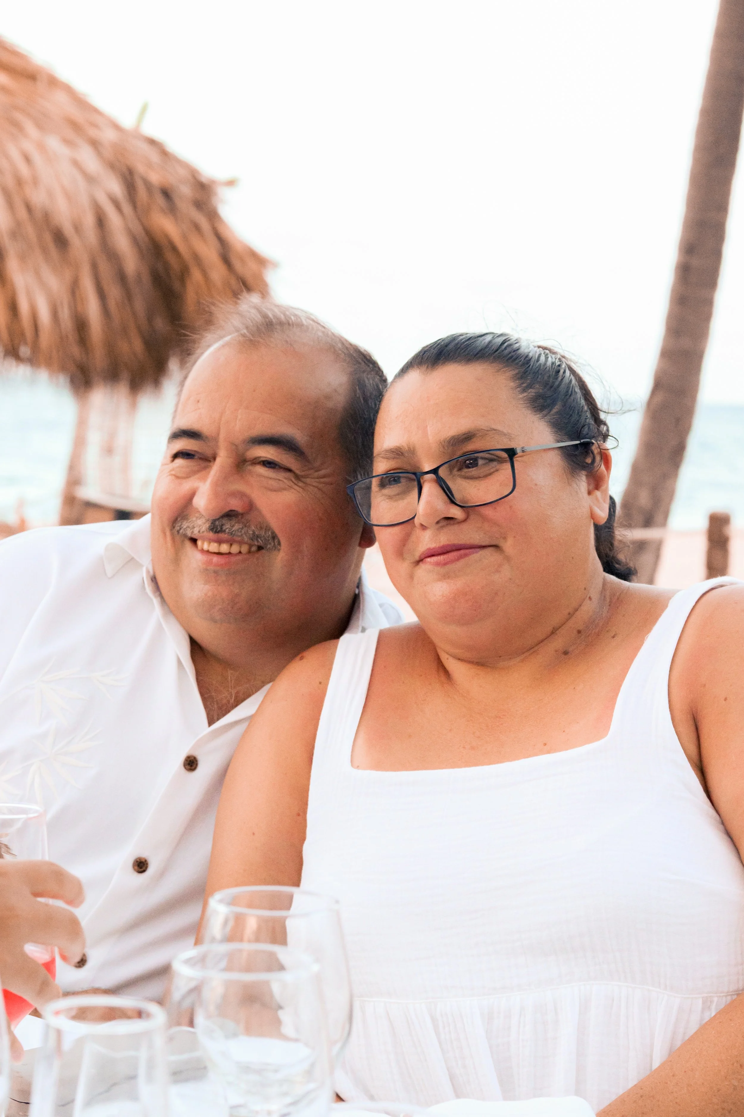 A smiling man and woman sitting close together at an outdoor beachside gathering, with glasses of water and a straw hat in the background.