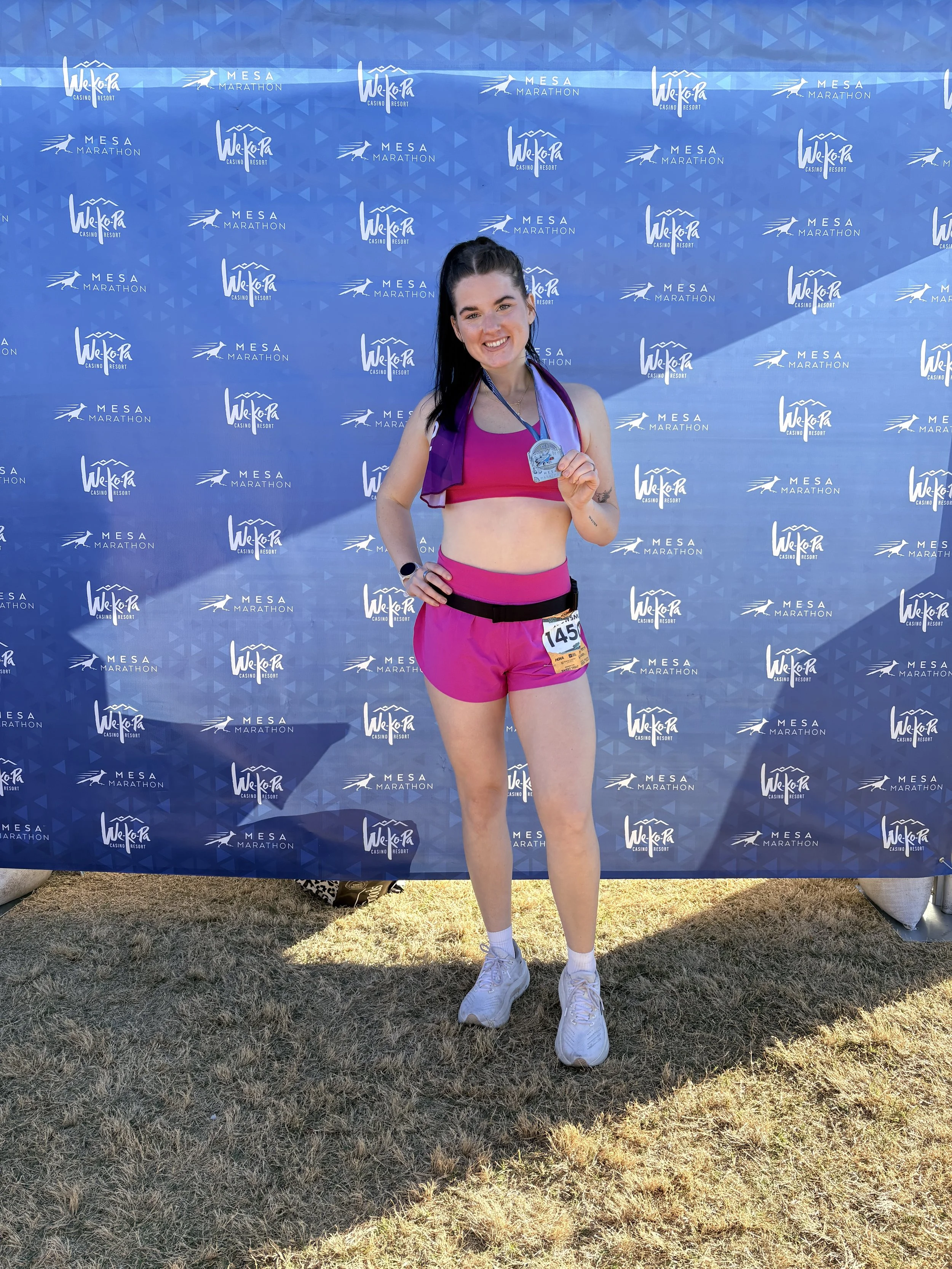 A young woman in pink running attire holding a medal at a marathon event, standing in front of a blue promotional backdrop with logos for Wek'a Casino Resort and Mesa Marathon.