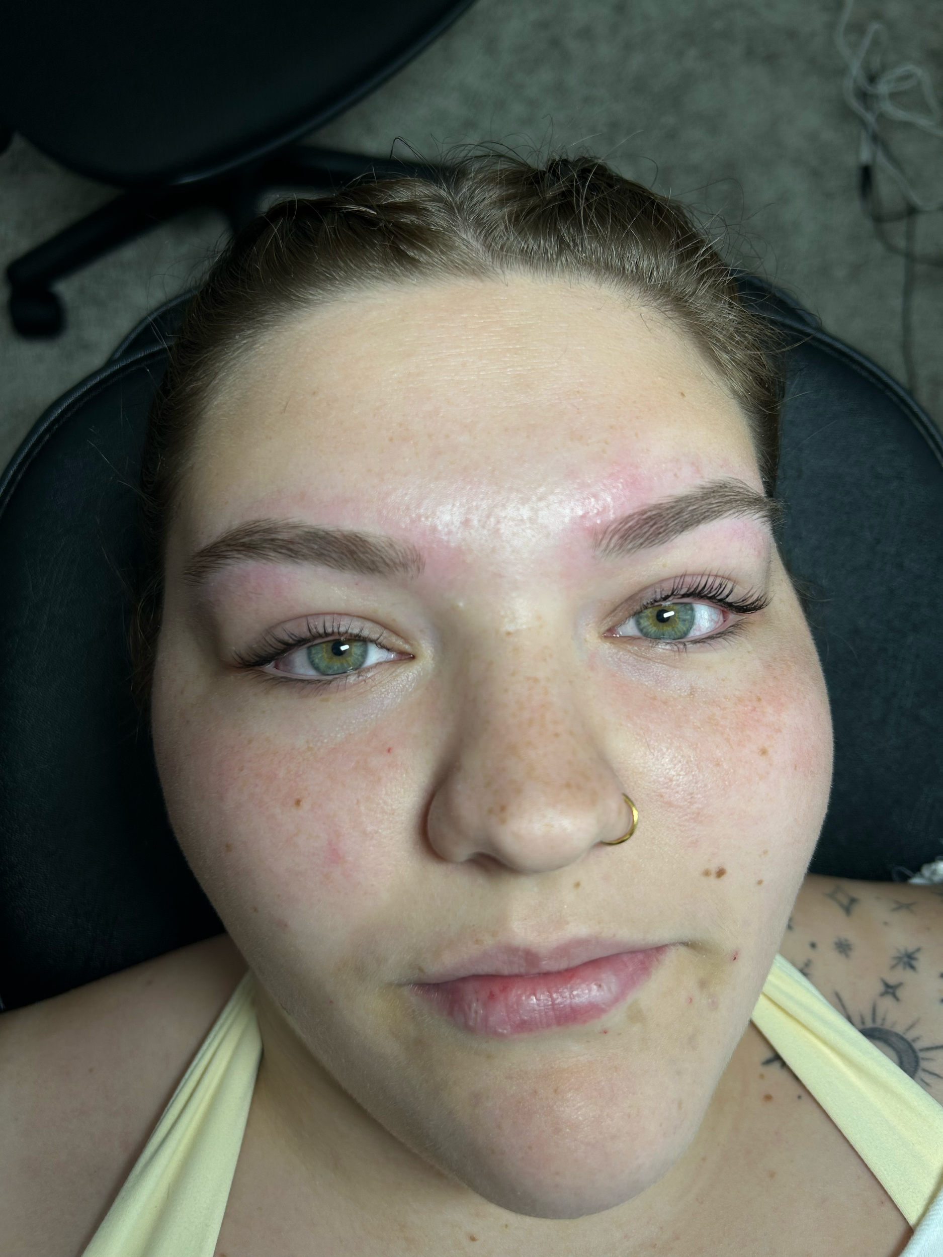 A woman lying on a chair with her eyes open, showing her face with freckles, a nose ring, and makeup on her eyelashes, in a room with a gray carpet and some wires in the background.