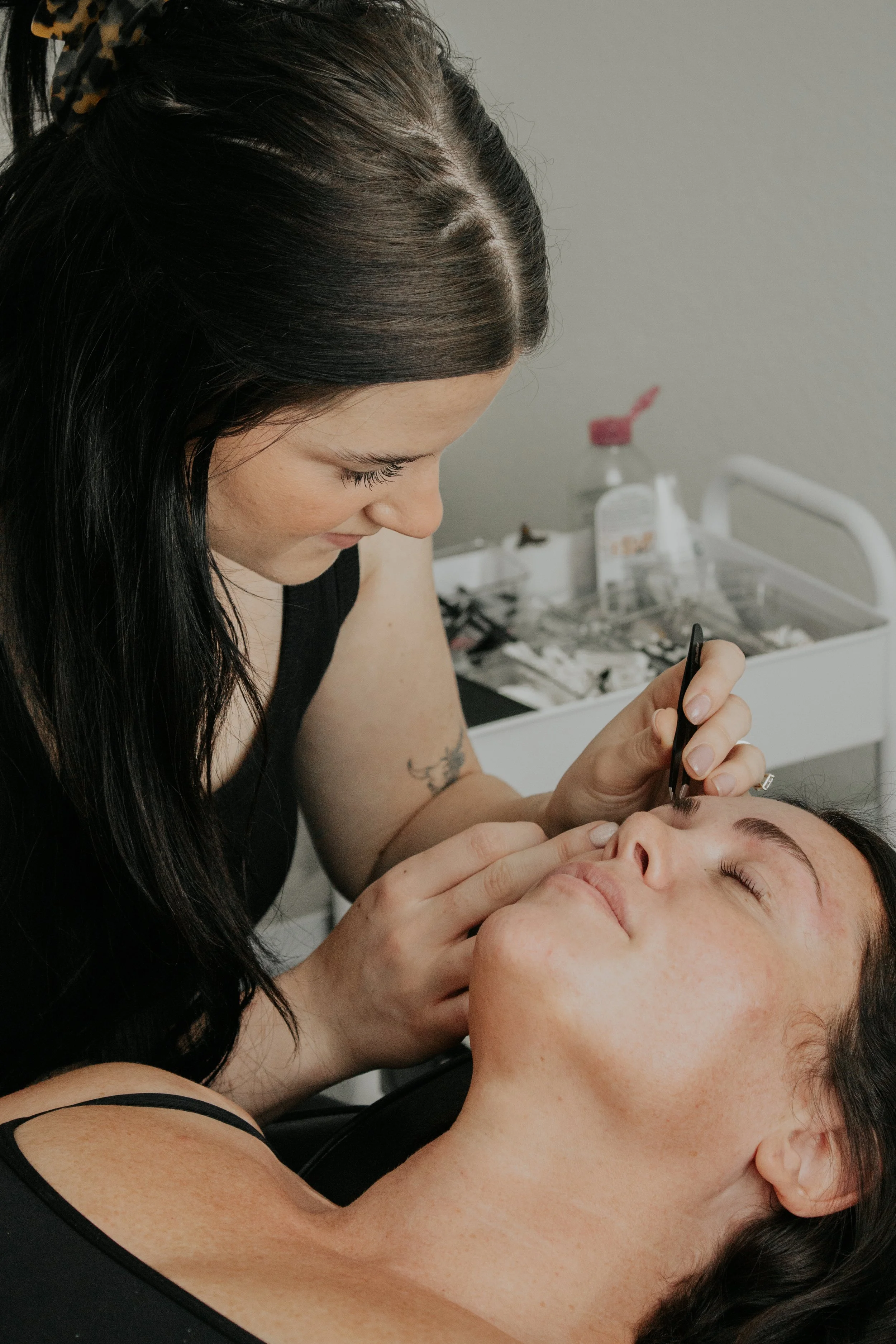 A woman with dark hair receiving a cosmetic procedure, lying back with her eyes closed, as another woman with dark hair and a black hairband uses a small tool on her face.