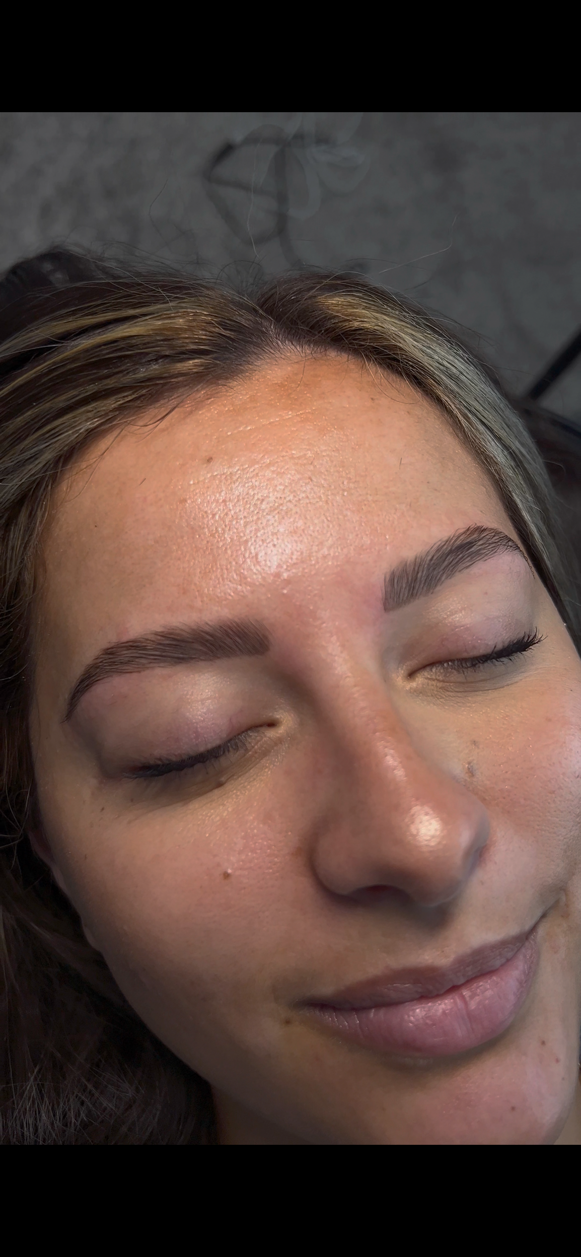 A woman with her eyes closed, showcasing freshly done eyebrow microblading tattooing in a professional setting.