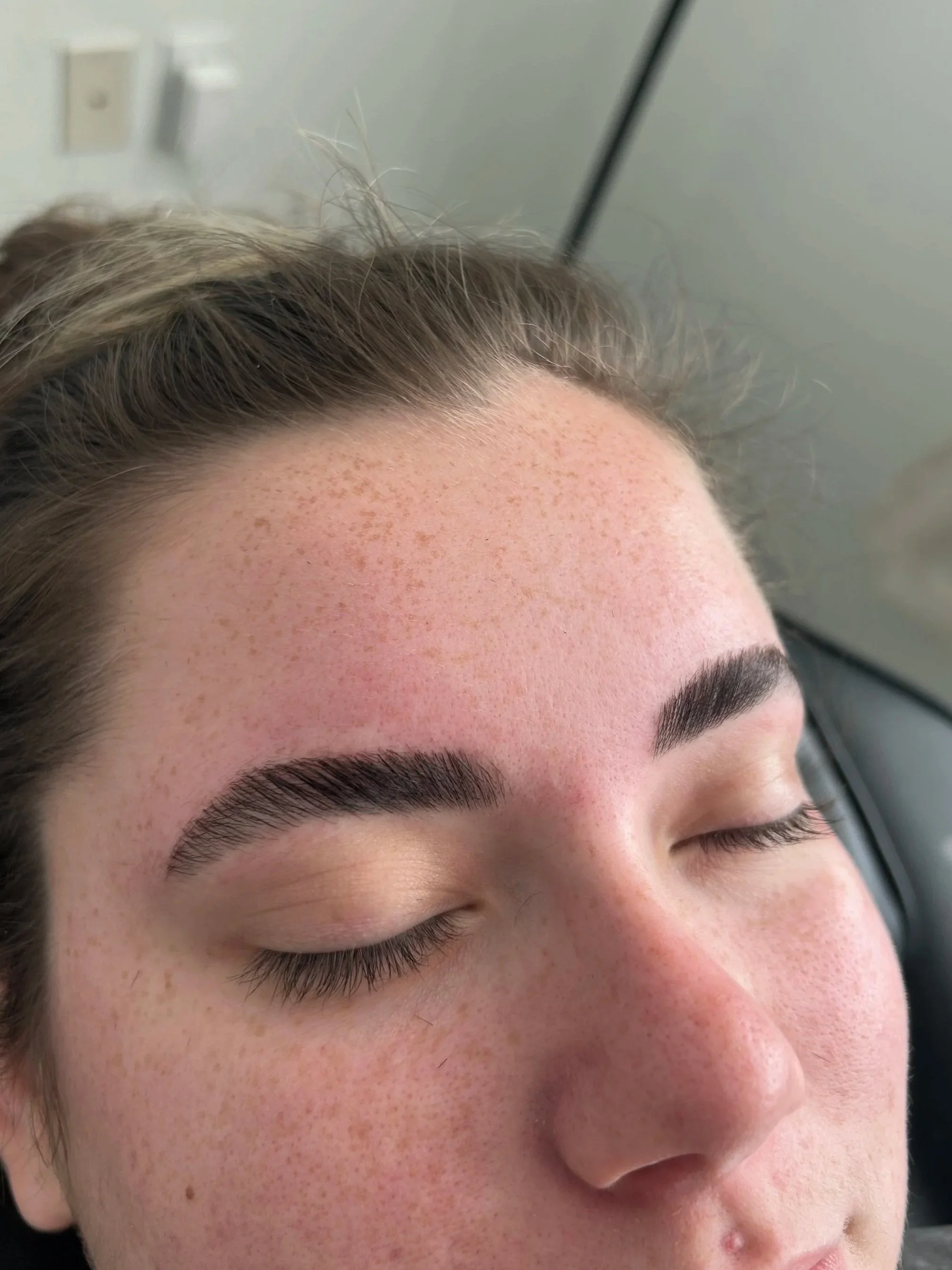Close-up of a person's face with freshly done dark, thick eyebrows and fair skin with freckles, resting with eyes closed.