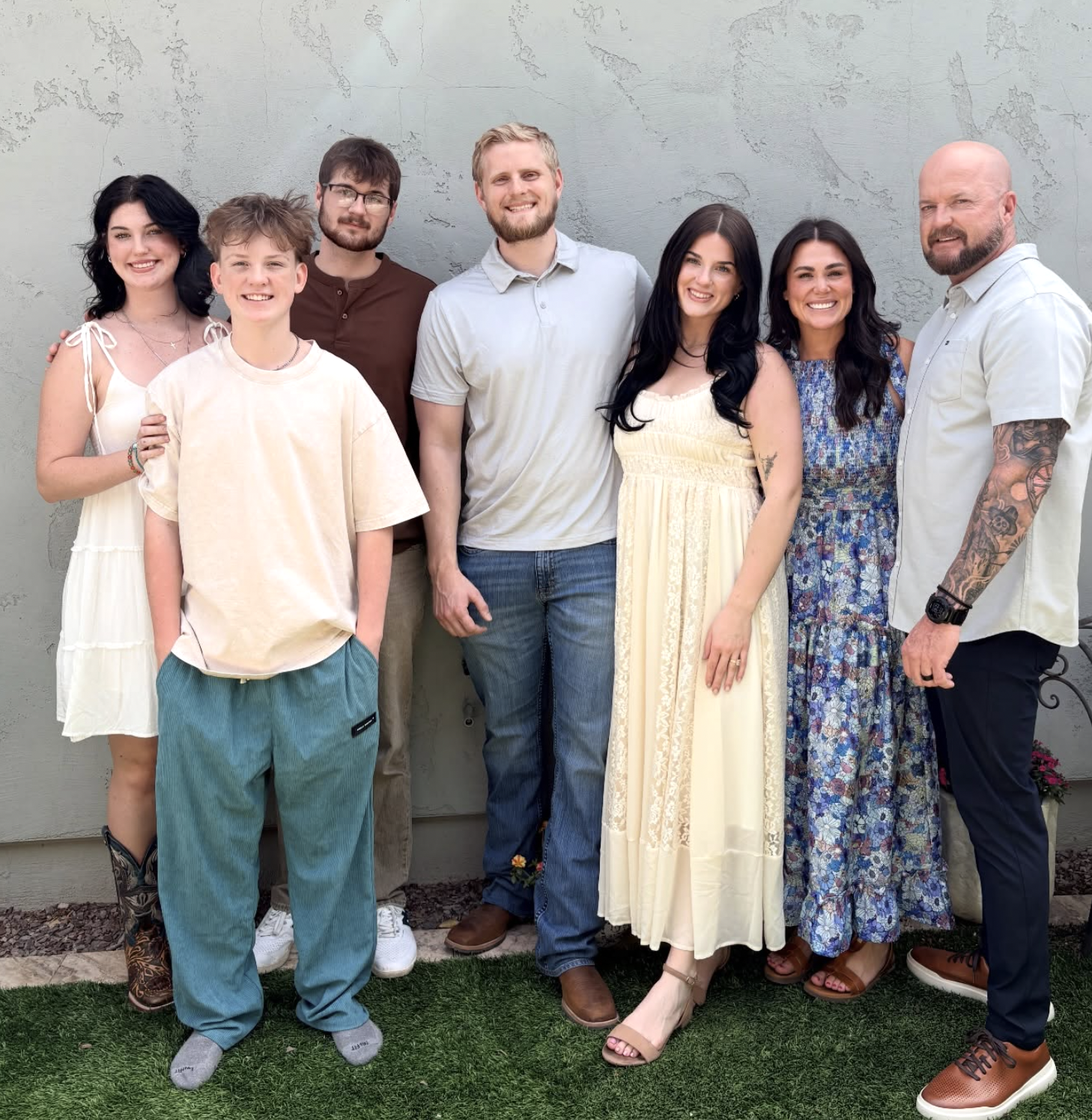 Group of eight people standing outdoors against a gray wall, smiling, dressed in casual and semi-formal summer attire.