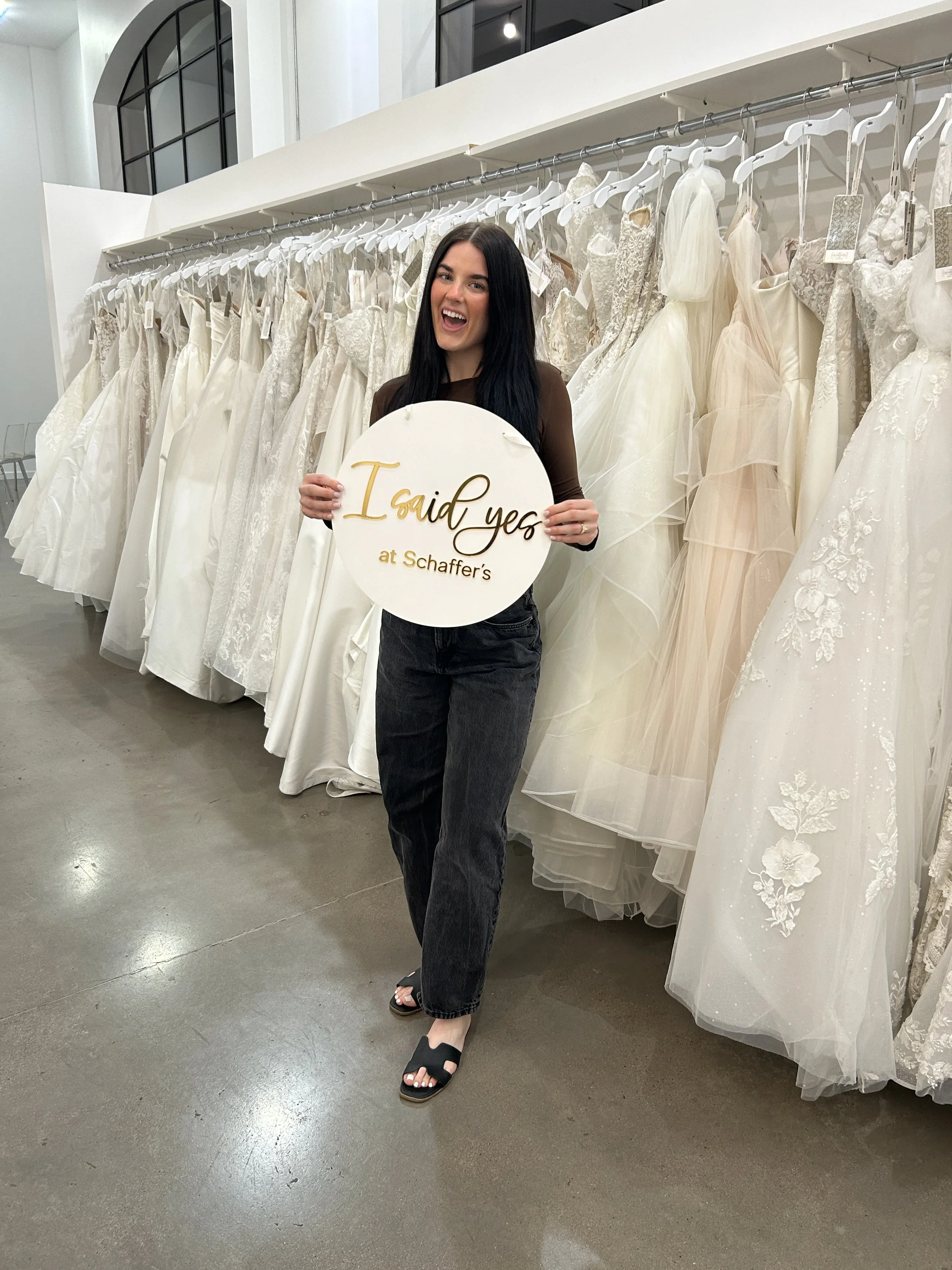 A woman standing in a wedding dress store, holding a sign that says 'I said yes at Schaffer’s', smiling.