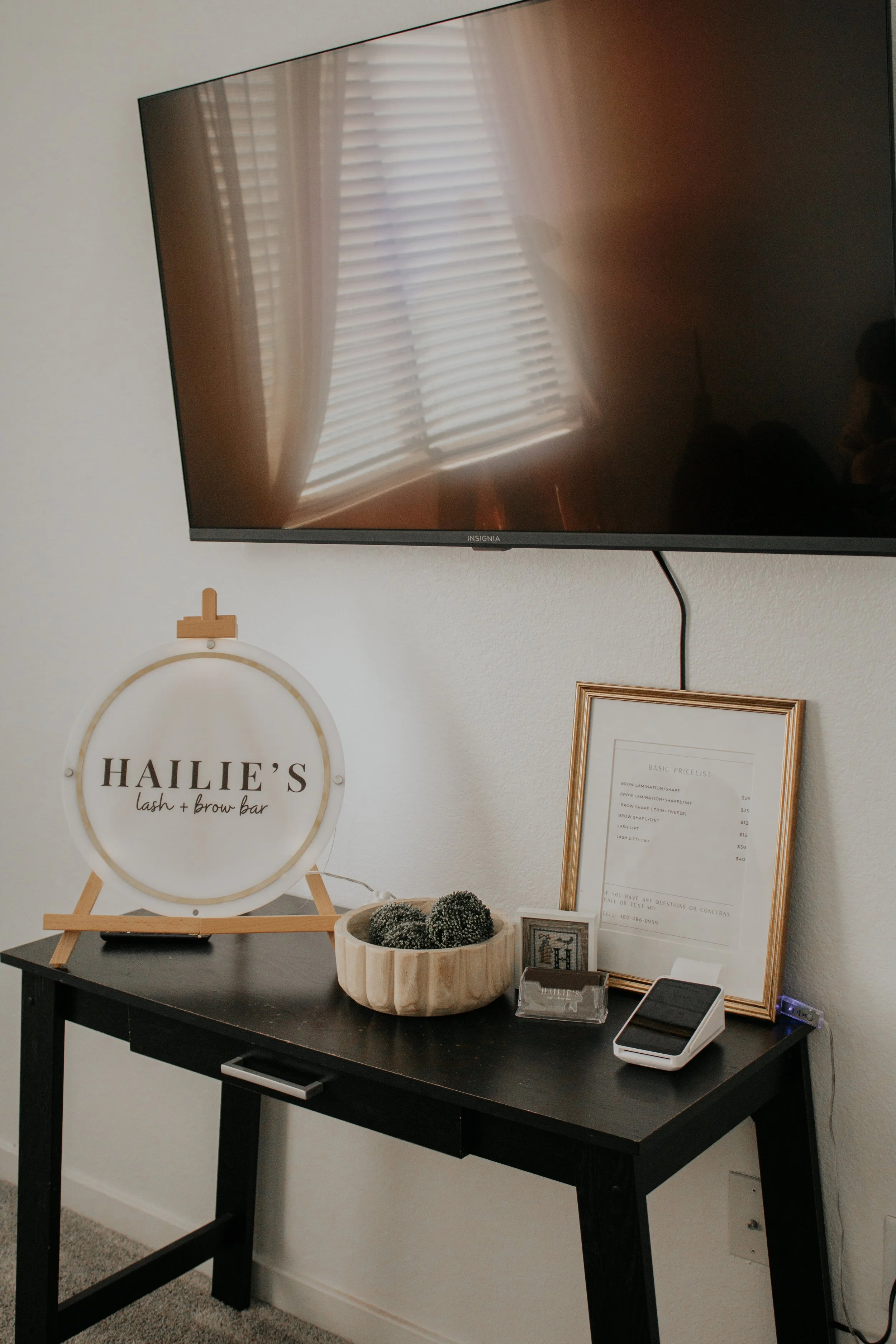 A black table against a white wall holding a framed sign, a bowl of decorative balls, a small framed picture, a white electronic device, and a large framed menu. A TV is mounted above the table reflecting curtains and a window.