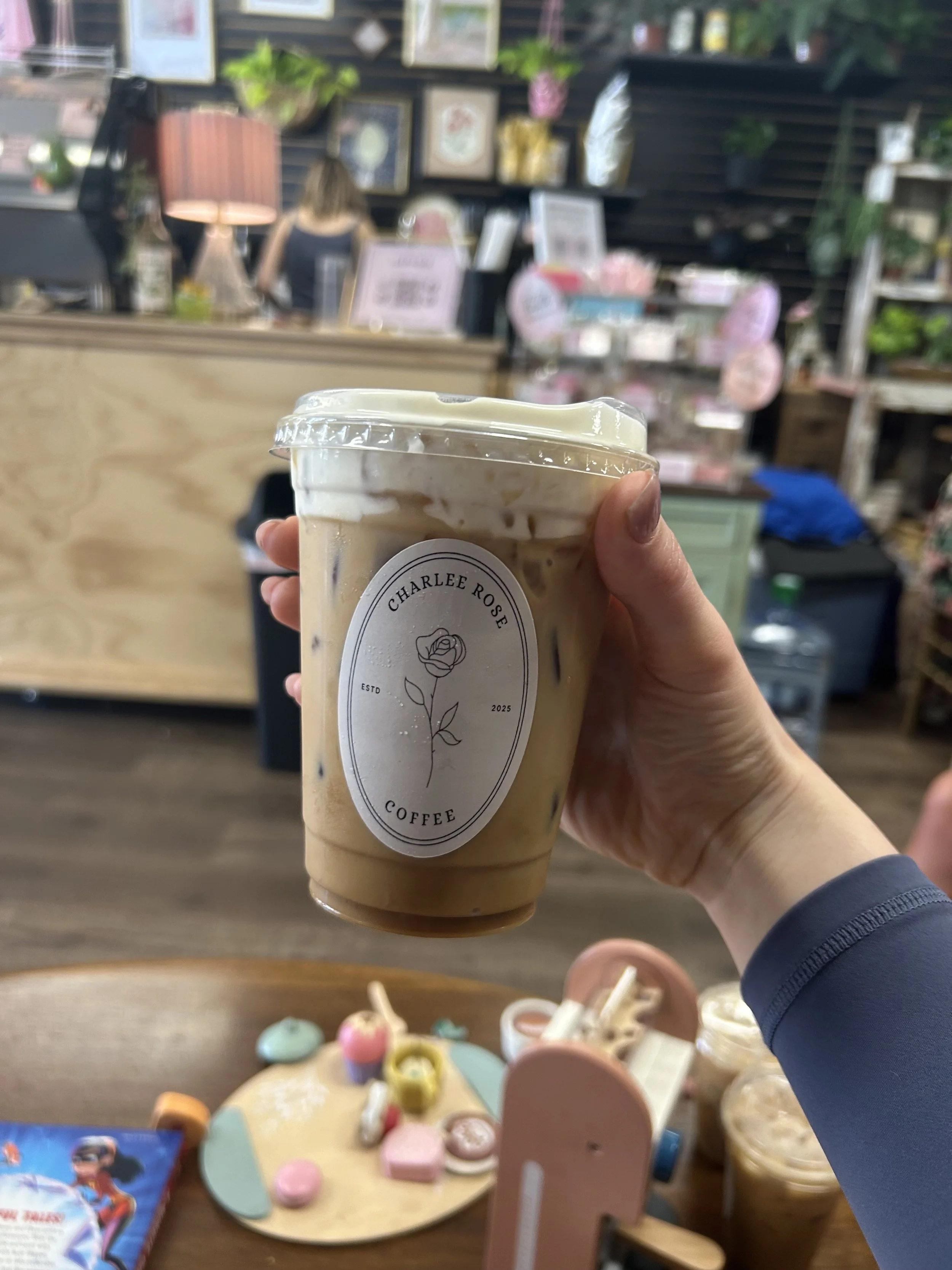 Hand holding a large iced coffee in a clear plastic cup with a charlee rose coffee sticker, inside a cozy, decorated coffee shop.