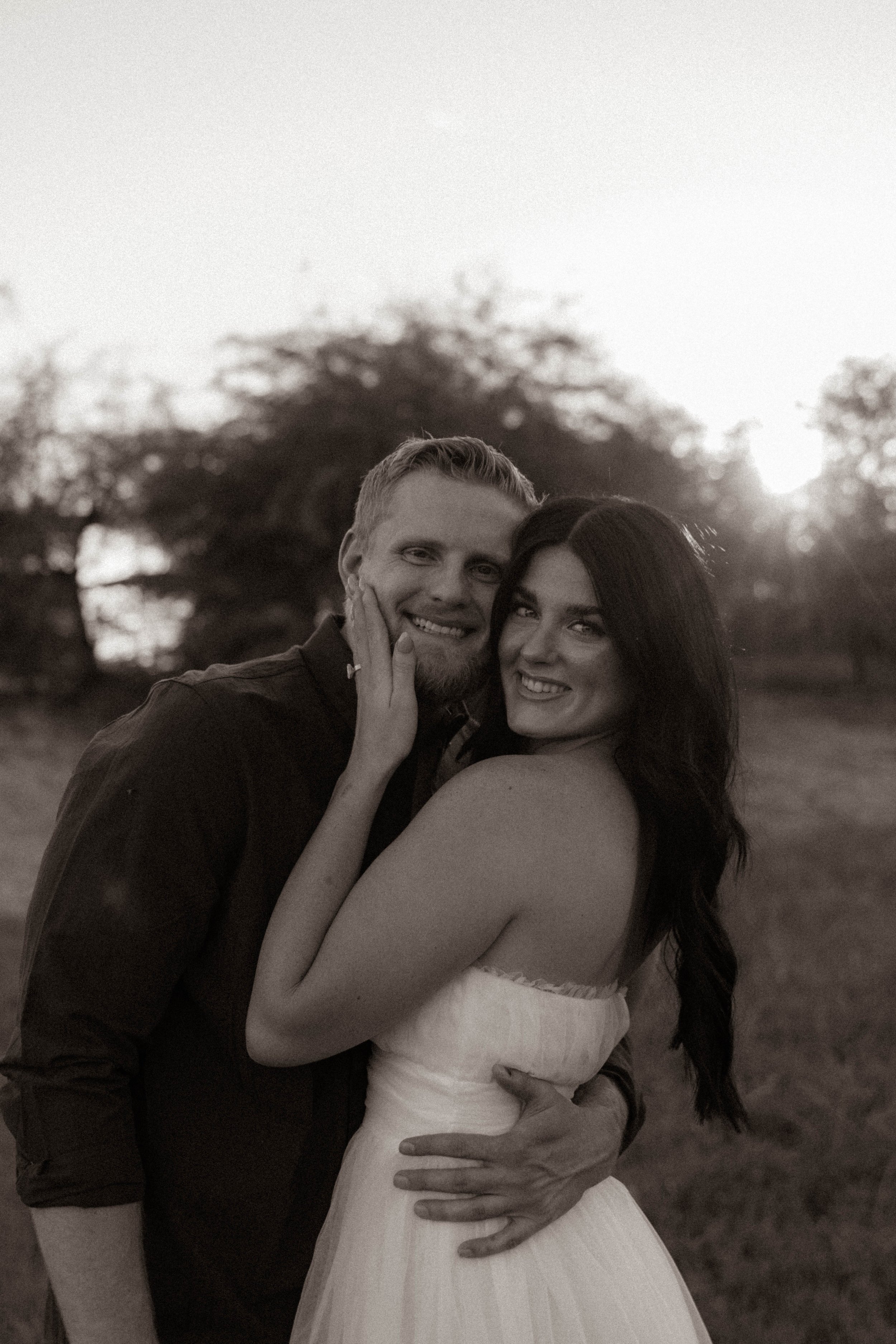 Black and white photo of a smiling man and woman embracing outdoors at sunset, with trees in the background.