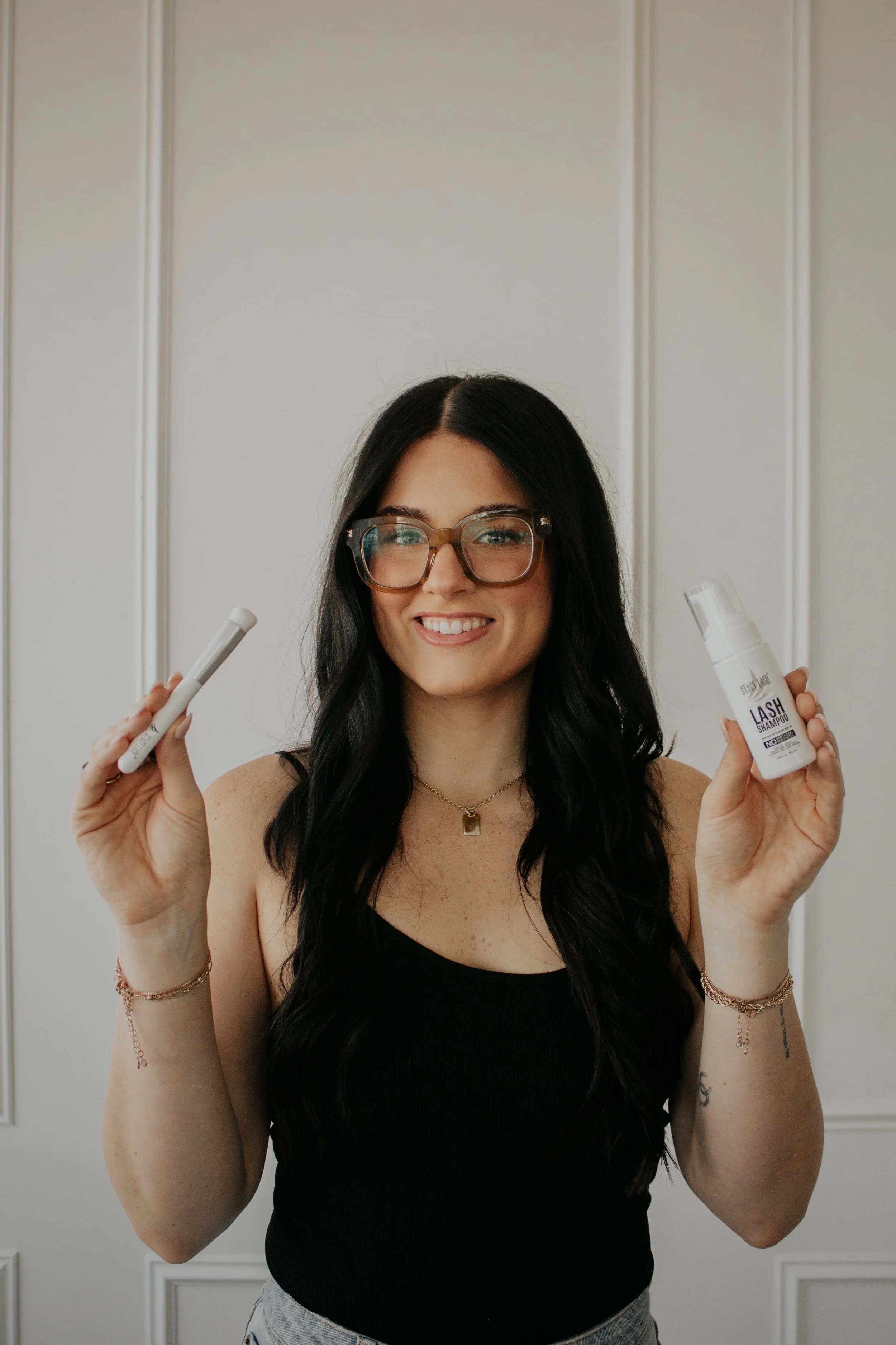 A smiling woman with long dark hair, glasses, and wearing a black sleeveless top holding eyelash glue and lash shampoo bottles, standing in front of a light-colored wall.