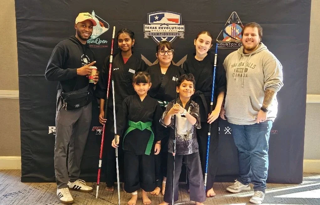 Group of seven people, including children and adults, standing in front of a backdrop with martial arts, holding martial arts weapons and wearing martial arts uniforms and casual clothing.